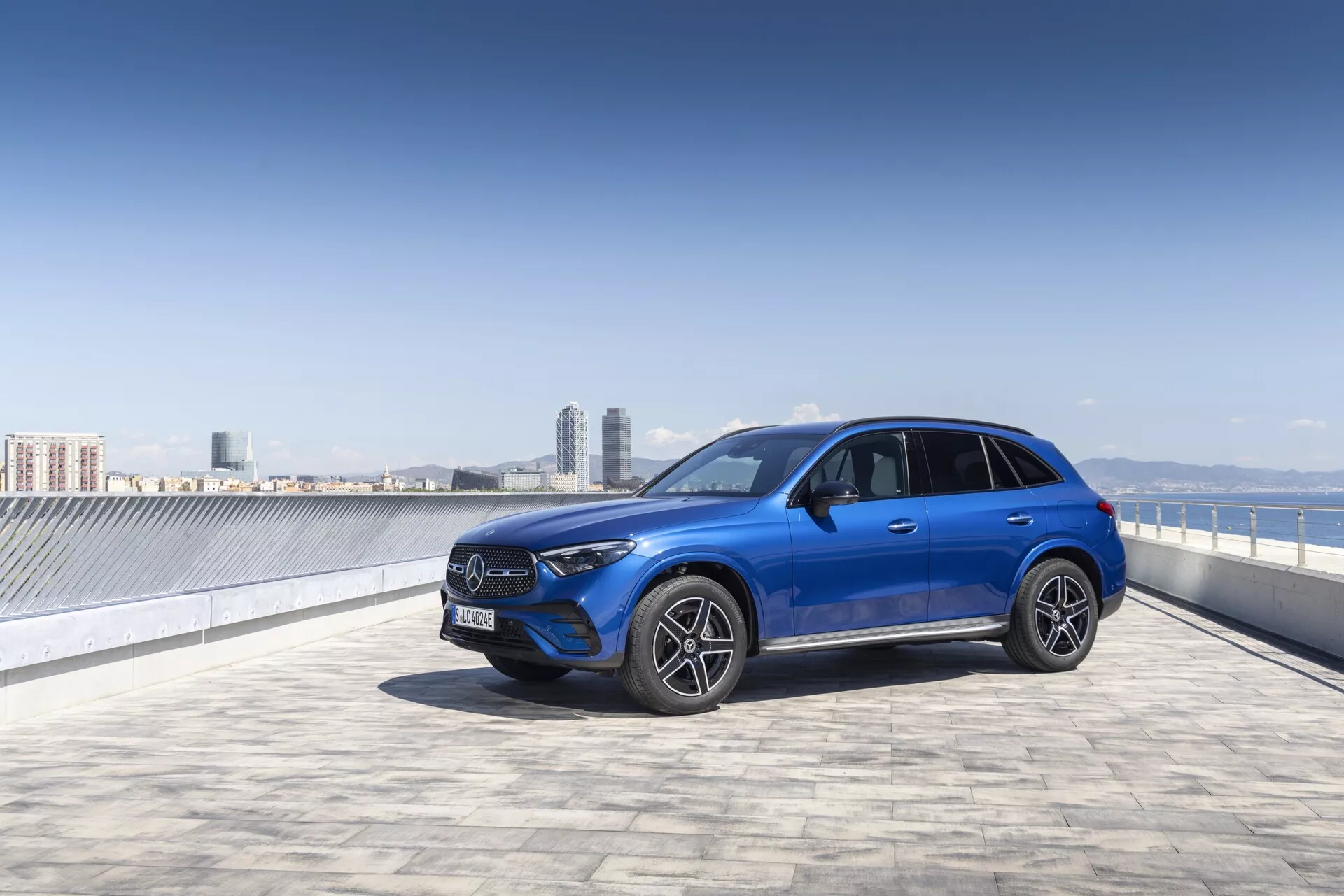 Ljusblå Mercedes GLC 2022 SUV på ett takparkeringsområde med klar himmel och stadslandskap i bakgrunden, fotograferad från en trekvarts främre vy. Bilden lyfter fram den framträdande frontgrillen med ett stort emblem, vinklade LED-strålkastare, främre stötfångare med möjliga sensorer, flerspokes legeringsfälgar och sidospeglar med integrerade blinkers. Bilens eleganta linjer och sportiga hållning är väl framträdande, vilket tyder på avancerad teknik och urban sofistikering.