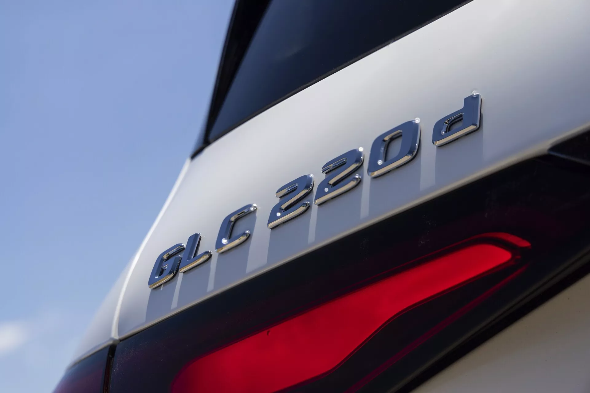 Närbild av bakdelen av Mercedes-Benz GLC 220d 2022, med särskilt fokus på modellbeteckningen precis ovanför den delvis synliga baklyktan. Bilden framhäver beteckningen 'GLC 220d', vilket antyder att det är en dieselvariant. Fotot från en låg, lätt uppåtvinklad vinkel visar de eleganta designlinjerna, den metalliska finishen på beteckningen som kontrasterar mot den blanka lacken, och de moderna LED-elementen i baklyktan, vilket tyder på avancerad belysningsteknik.