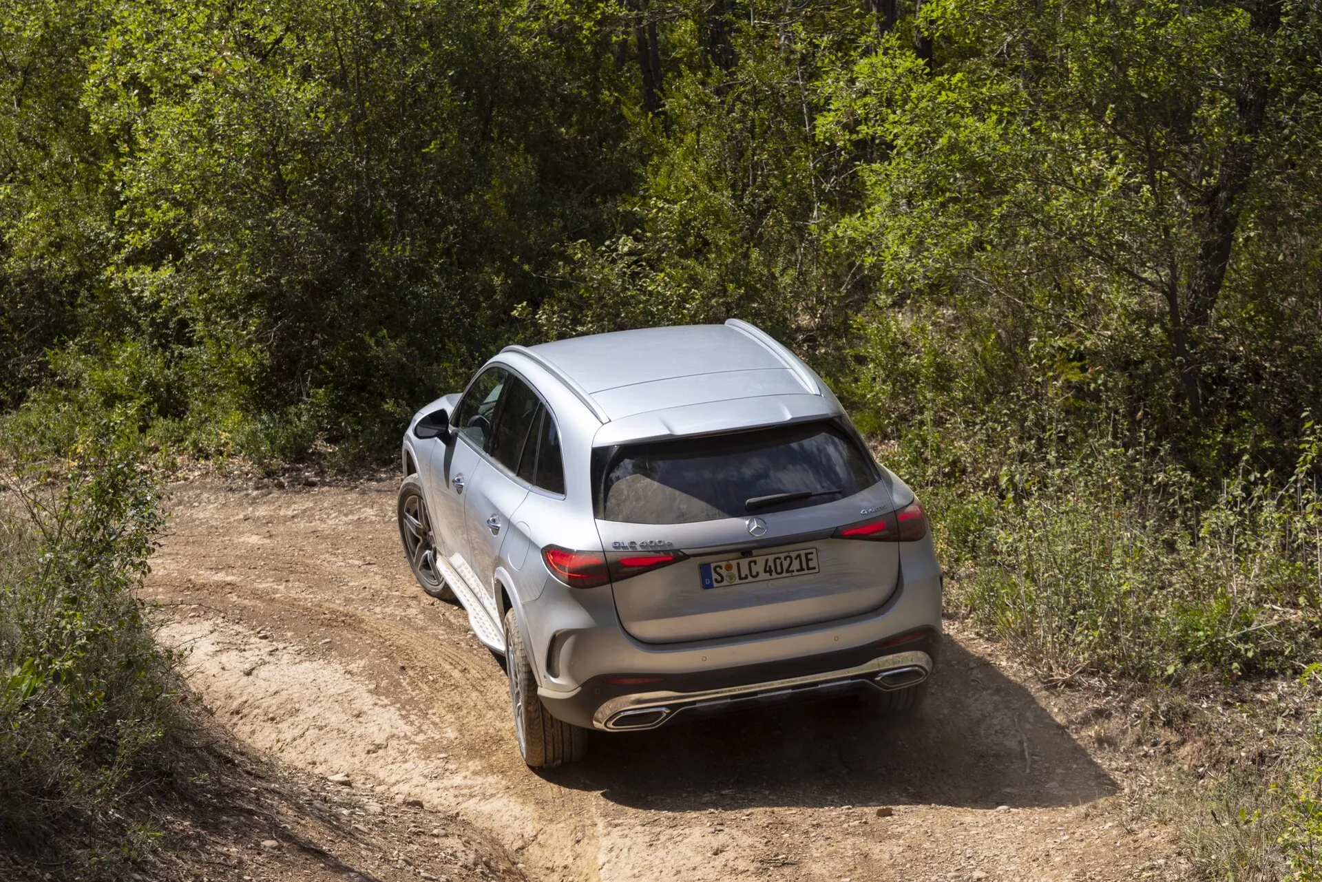 Bilden visar en modern, lyxig crossover eller SUV som tagits från en upphöjd, bakre trekvartsvinkel, vilket tydligt visar bilens bakre och delvis dess sidoprofil. Fordonet har skarpa, horisontellt orienterade baklyktor, stängt baklucka, synliga bakre vindrutetorkare och en bakre stötfångare med integrerade avgasrör. Det synliga bakre legeringshjulet har en flerspokesdesign med däck av måttlig profil, lämpliga för lätt terrängkörning. Bilen, i silver eller ljusgrå metallisk färg, har ett panoramasoltak och monterade takräcken. Den står stilla på en smal, grusväg omgiven av gröna löv, vilket antyder en äventyrlig, utomhus livsstil.