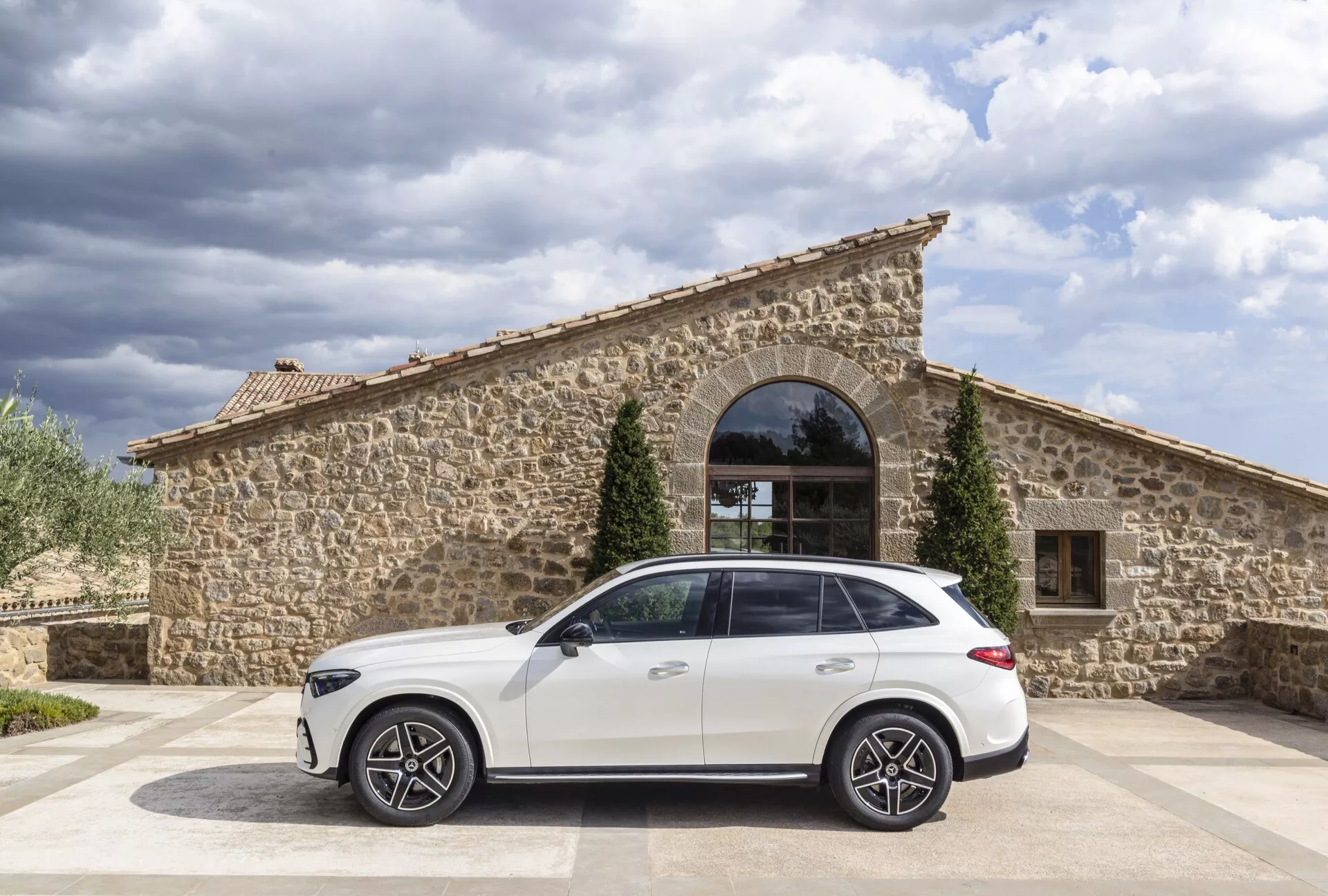 Sido vy av en vit 2022 Mercedes GLC SUV parkerad på en kullerstensyta med en rustik stenbyggnad med ett stort bågformat fönster i bakgrunden. Den mulna himlen ger naturligt dagsljus, och kameravinkeln visar bilens tak och karosslinjer från passagerarsidan. Noterbara funktioner inkluderar stora flerspåkiga aluminiumhjul, en karaktärslinje under fönstren, infällda dörrhandtag, takräcken och en hajfenantenn.