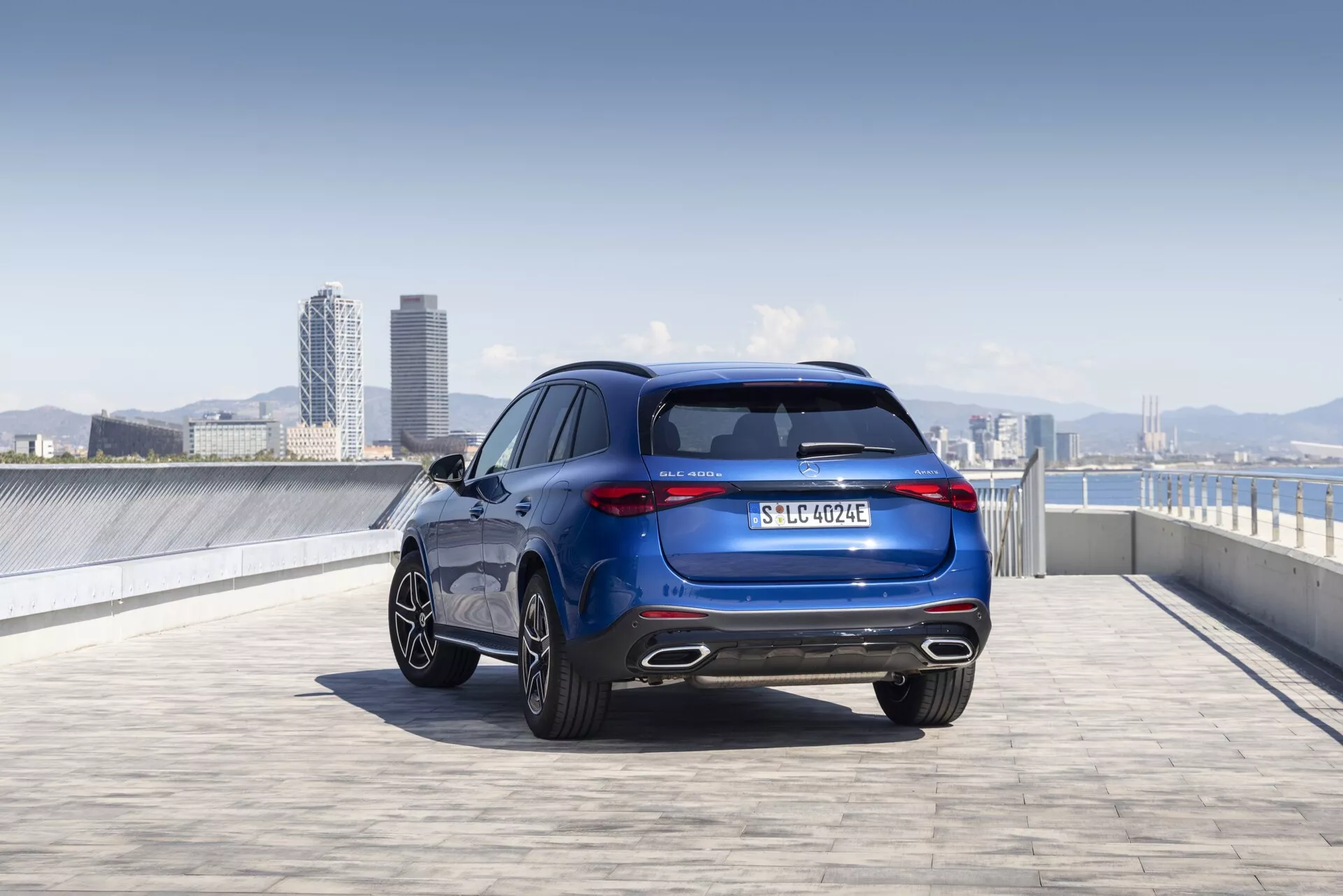 Lågvinkelbild av en 2022 Mercedes GLC (254) från en bakre trekvartsvy som visar upp dess eleganta och sportiga designlinjer, bakljus, aluminiumfälgar, metalliclack och märkning. Bilden fångar hela fordonets höjd och bredd, med betoning på detaljer som bakrutan, avgasystemet, registreringsskylten och eventuellt en emblem för utrustningsnivå eller drivlina. Den utomhusmiljön, troligtvis en takparkering med en stadssilhuett i bakgrunden, framhäver fordonets moderna och sofistikerade attraktionskraft.