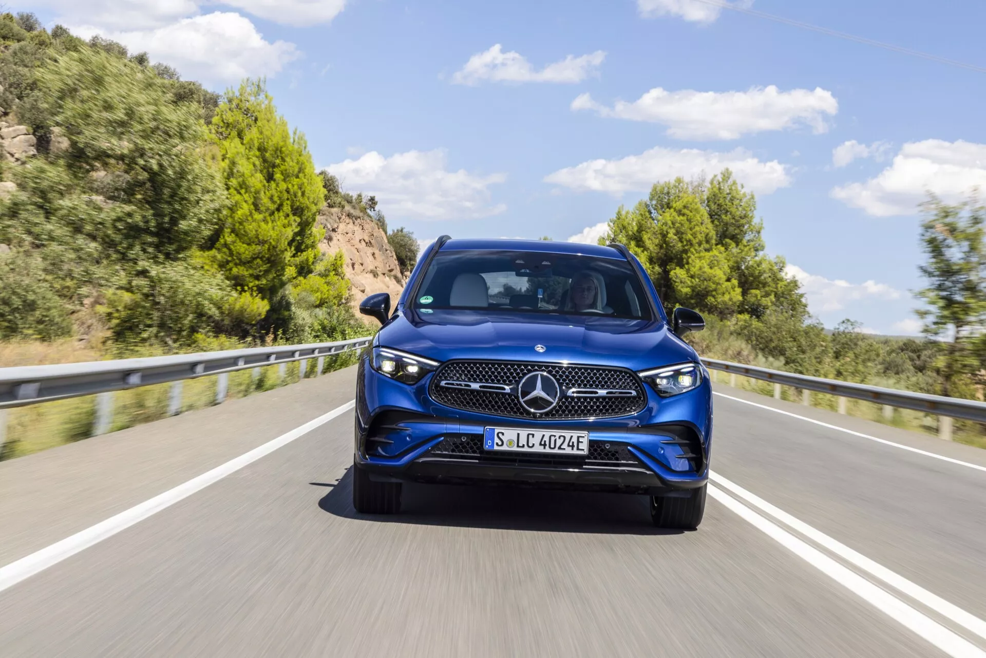 Bild av en blå 2022 Mercedes-Benz GLC 254-modell fotograferad från en främre trekvartsvinkel som framhäver den eleganta designen, det framträdande treuddiga stjärnemblemet, det detaljerade galler-mönstret, de eleganta strålkastarna som smälter in i stänkskärmarna och tydliga detaljer av den främre stötfångaren, motorhuven och dörrarna. Bilen, fotograferad i rörelse på en naturskön tvåfilig väg med frodiga kullar i bakgrunden, har troligen elektrisk eller hybridteknik som föreslås av 'EQ'-märkningen på registreringsskylthållaren. Bilden är detaljerad med rörelseoskärpa som indikerar rörelse och visar bilens dagsljus under dagtid med klar himmel.
