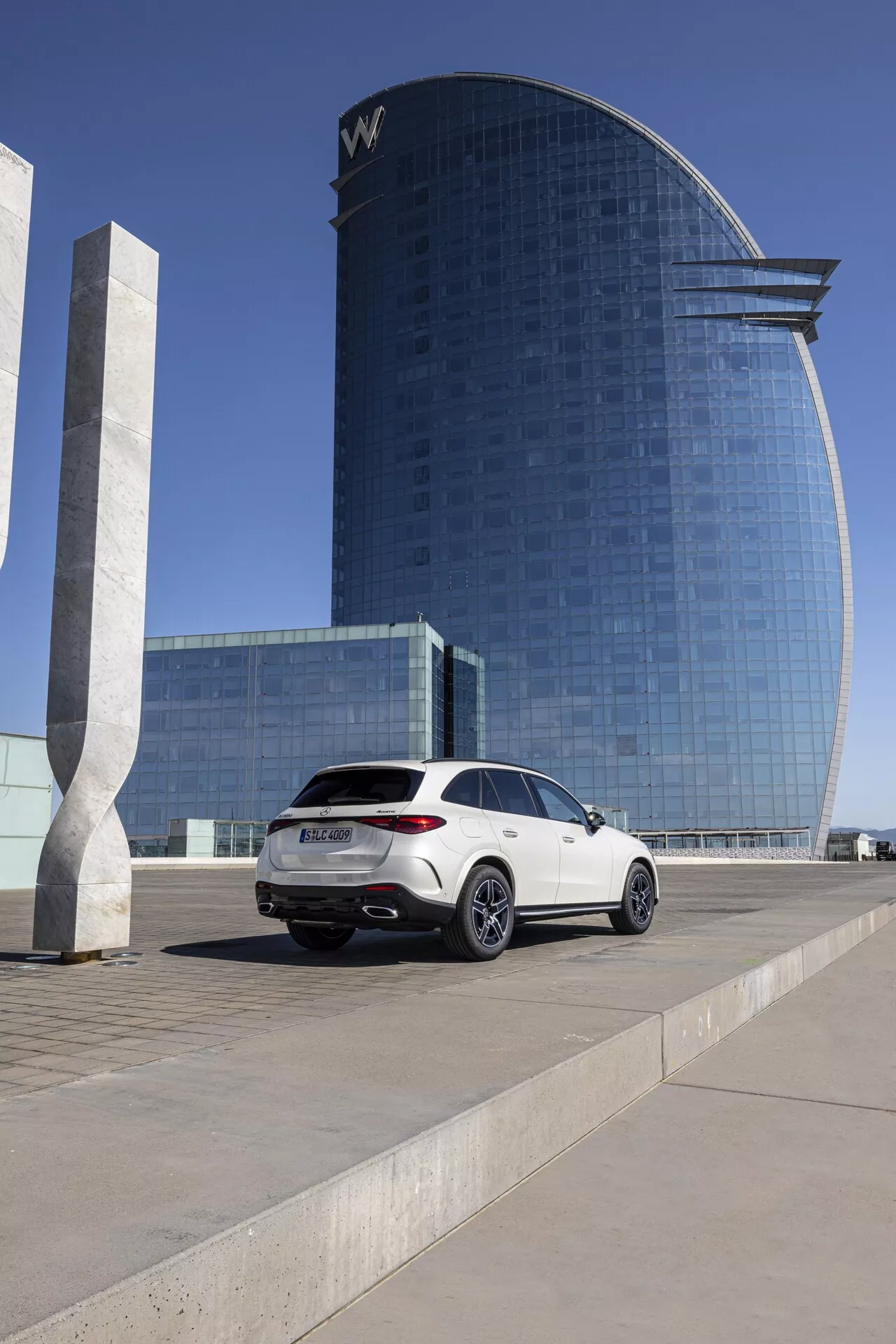 Mercedes GLC 2022 - Vit lyxig SUV parkerad på ett torg; medellåg vinkel som fångar passagerarsidans profil och bakparti, visar moderna bakljus, dubbla avgasrör, flerspoked fälgar, coupé-liknande taklinje, tonade rutor; bakgrund med modern hotell- eller kontorsbyggnad med reflekterande glasfasad i starkt dagsljus.