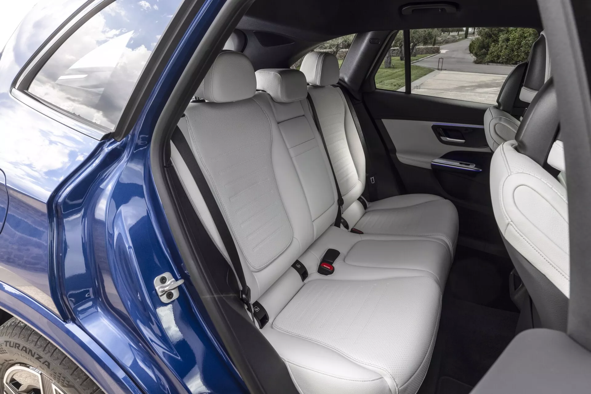 Bild av interiören på Mercedes GLC 2022 som visar det bakre passagerutrymmet sett genom höger bakdörr. De ljusa, läderklädda baksätena har nackstöd och ett fällbart armstöd med mugghållare. Bakdörren innehåller en högtalarenhet, elhissar och en förvaringsficka. Framsätenas ryggstöd har förvaringsfickor och det rymliga bakre området framhäver bilens familjevänliga design och lyxiga funktioner med naturligt dagsljus som lyser upp scenen.