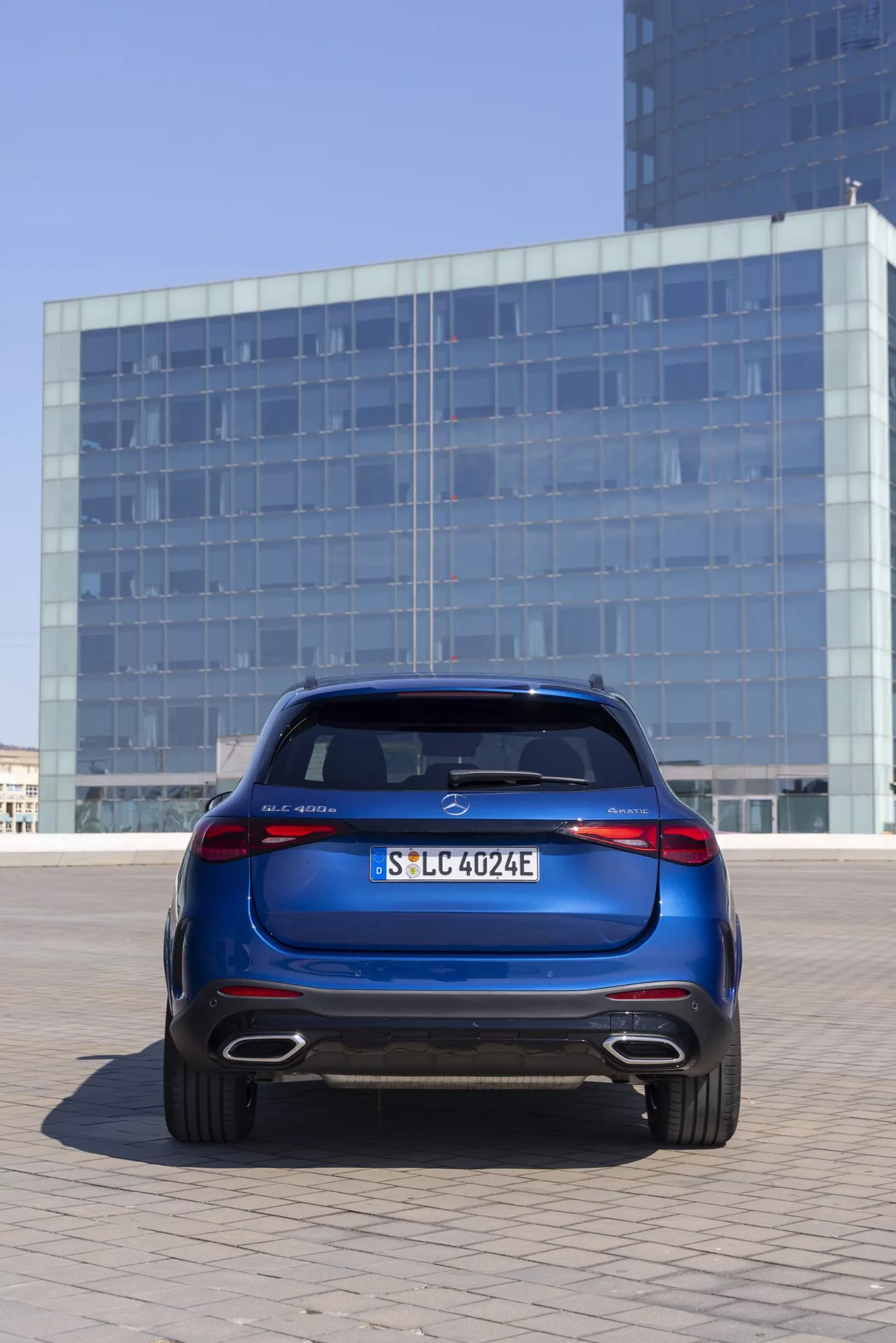 Bakre vy av 2022 Mercedes GLC (254) i en djup metallicblå färg, placerad utomhus i en urban miljö med en byggnad med reflekterande glasfasad i bakgrunden. Bilen är centrerad, fångad från en något låg vinkel som framhäver bakluckan, bakre stötfångaren, LED-bakljus, lättmetallfälgar och takspoiler. Funktioner inkluderar ett framträdande centeremblem, registreringsskylt, modellbeteckning på vänster sida, 'E' som indikerar en elektrisk eller hybrid modell på höger sida och en bakre diffuser med avgasuttag, vilket antyder att det är en hybridmodell.