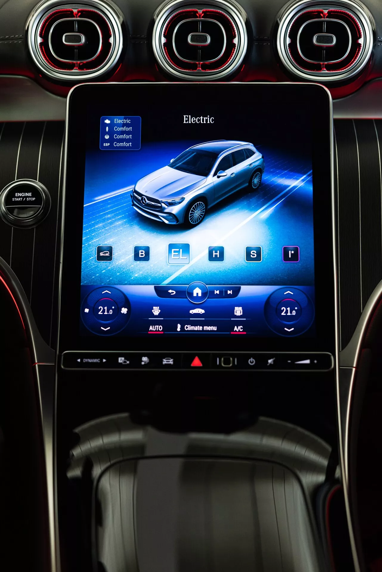 Interiörvy av en bils instrumentbräda med Mercedes GLC 2022 infotainmentsystem. Fokuspunkten är en vertikalt orienterad pekskärm inbäddad i instrumentbrädan. Det livfulla gränssnittet visar en grafisk avbildning av ett blått elfordon i en lätt 3/4 främre perspektiv. Ordet 'Electric' visas högst upp på skärmen, vilket indikerar fordonets drivlina typ. Skärmen innehåller också inställningar för tvåzoners klimatkontroll med en temperaturinställning på '21' på båda sidor, tillsammans med plus- och minusjusteringsknappar och olika ikoner längst ner. Materialen på instrumentbrädan runt skärmen verkar inkludera glansiga och texturerade plaster samt eventuellt läderdetaljer. Ovanför skärmen finns runda ventilationshål med röda accenter, vilket ger bilens interiör en sportig eller exklusiv estetik.