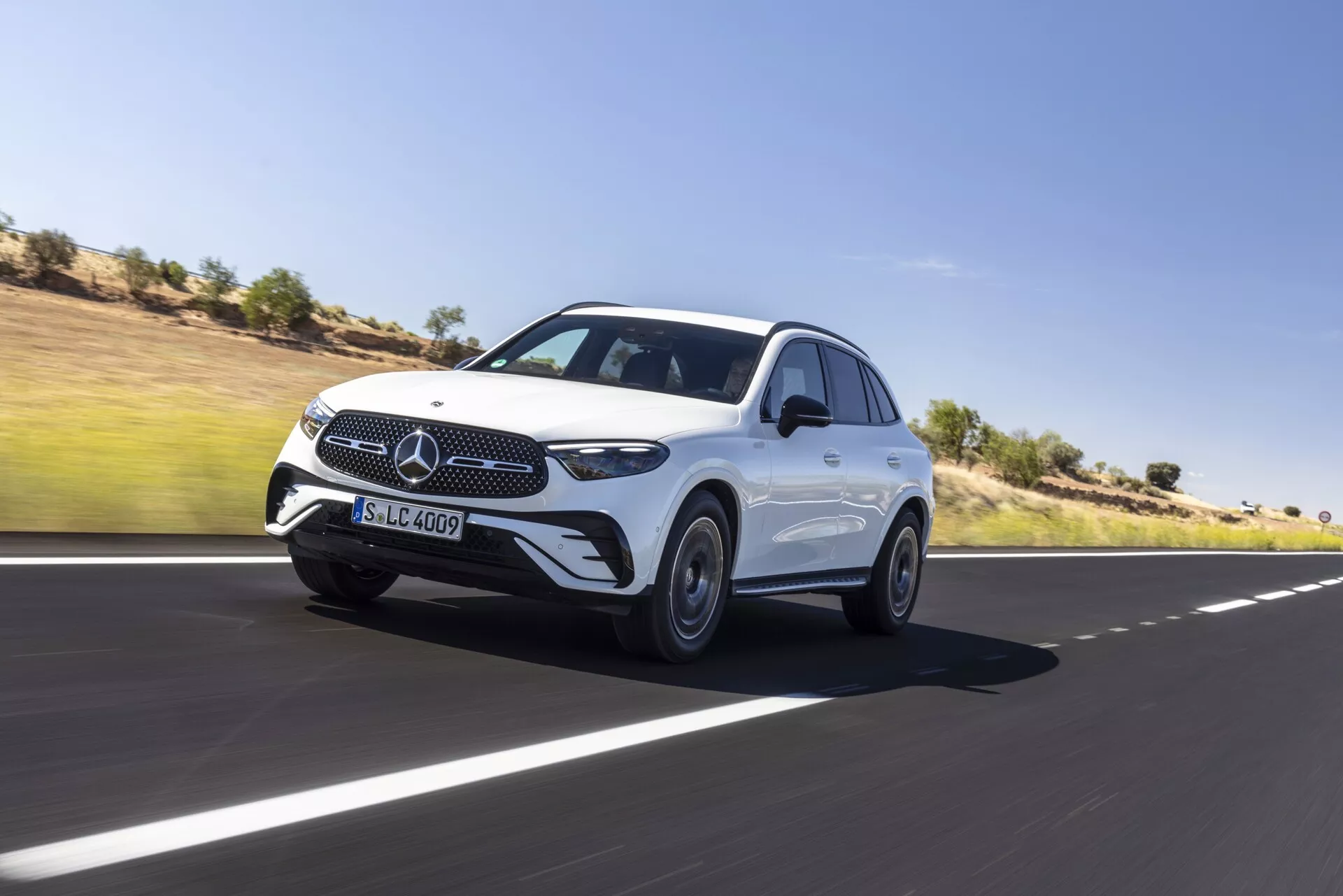 Dynamisk bild av lyx-SUV:n Mercedes GLC 2022 som kör framåt på en slät asfaltsväg, som visar dess fram- och sidoprofil. Funktioner inkluderar den ikoniska grillen med trefärgad stjärna, eleganta LED-strålkastare, sensorer på främre stötfångaren som indikerar avancerade förarassistanssystem, skulpterade huvlins, lättmetallfälgar, strömlinjeformade sidobackspeglar med integrerade blinkers och en flödande taklinje för en sportigare siluett. Fordonets dominerande hållning och lätt rörelseoskärpa runt hjulen betonar dess rörelse. Bakgrund med klar himmel och ökenlandskap.