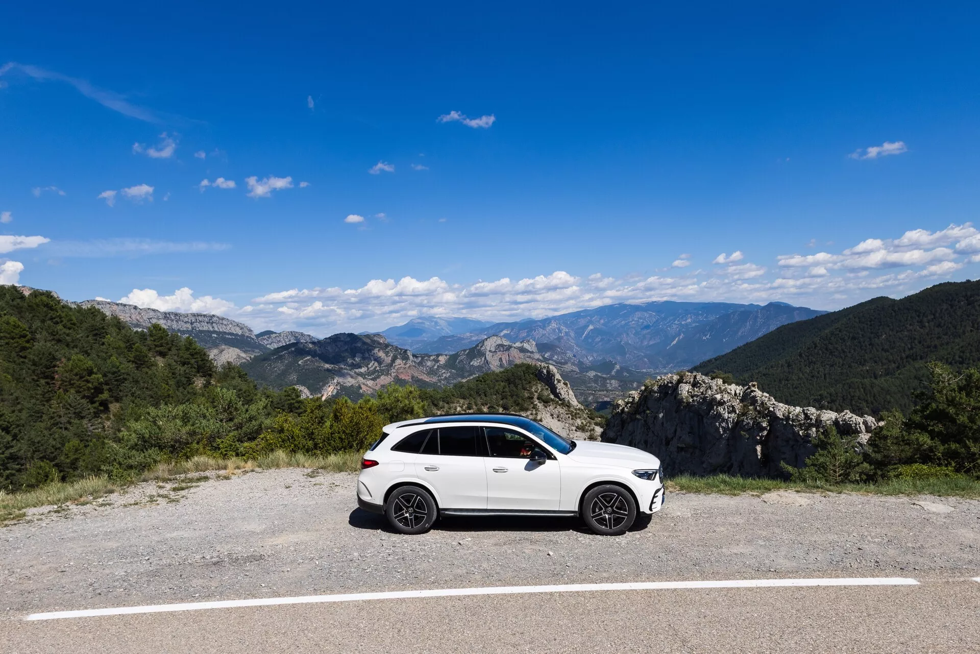En vit, sportig Mercedes GLC SUV från 2022 står parkerad på ett plant, asfalterat område omgiven av ett naturligt bergslandskap under en klarblå himmel. Sidoprofilen visar fordonets förarsida och framhäver designelement som legeringshjul, sidospeglar, fönsterlister och karosslinjer. Belysningen och klarheten låter de subtila konturerna på sidodörrarna vara synliga.