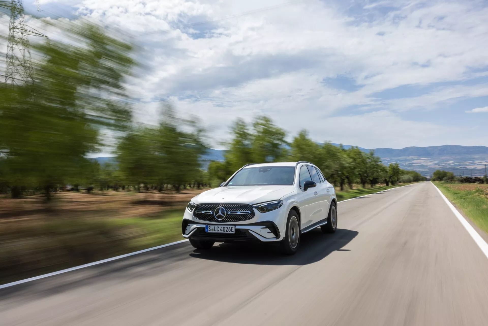 Främre trekvarts vy av en vit Mercedes GLC 2022 i rörelse på en tvåfilig väg, som visar dess främre grill, strålkastare, del av huven, vindrutan och förarsidan. Bakgrunden är rörelseoskärpt, vilket framhäver bilens hastighet. Den eleganta designen, den stängda grillen, energieffektiva LED-strålkastare och synliga dagskörljus, tillsammans med ett modernt landskap med suddiga träd och kuperad terräng, presenterar ett dynamiskt och stiligt utseende som är idealiskt för reklammaterial.