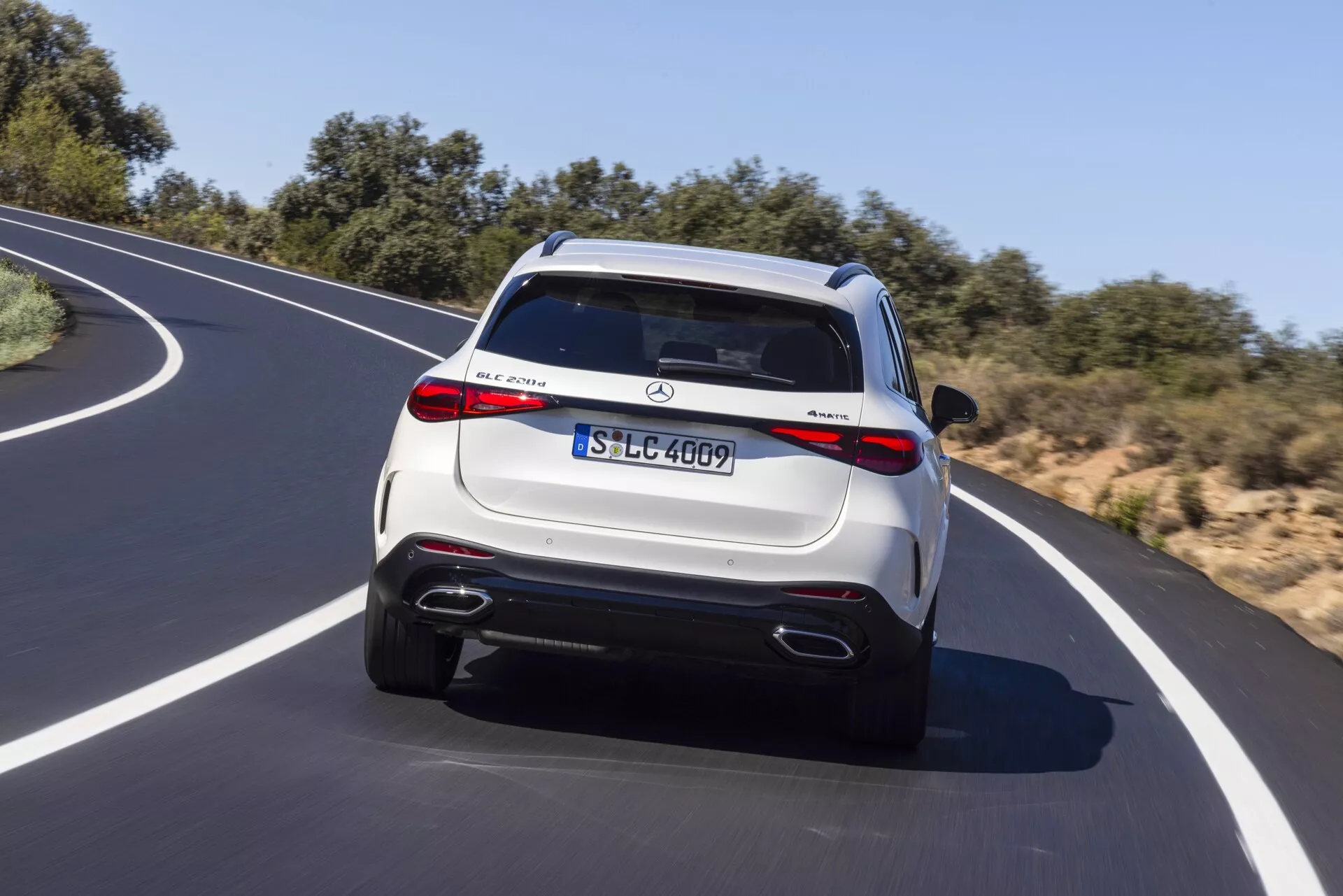 Bakrevy av en rörlig Mercedes-Benz GLC 2022 SUV på en kurvig asfaltväg under en klar himmel. Fordonet har en ljus metallisk finish, LED-bakljus, dubbla avgasrör, en bakre stötfångare med diffusor-liknande element och en takspoiler med integrerat bromsljus. Synliga emblem och en europeisk registreringsskylt indikerar modell- och trimspecifikationer. Mångekrade fälgar och potentiella högteknologiska funktioner som parkeringssensorer och en backkamera tillför en lyxig känsla.