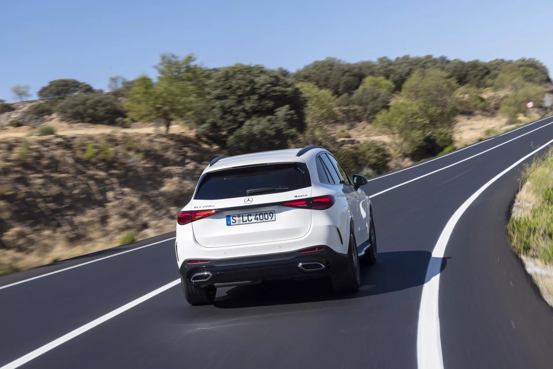 Bakre vy av en modern Mercedes GLC SUV 2022 i rörelse, som visar dubbla avgasrör, aerodynamisk bakre diffuser, LED-bakljus, sportig bakspoiler och praktiska funktioner som bakre vindrutetorkare och högt monterat tredje bromsljus. Färgen på SUV:n är ljus, som vit eller silver, vilket förhöjer dess lyxiga utseende medan den kör på en slät asfaltväg omgiven av naturlig växtlighet. Rörelseoskärpa framhäver fordonets dynamiska prestanda.