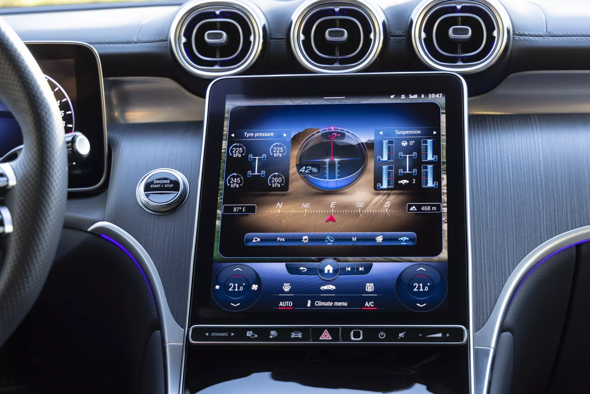 Närbild av interiören i en Mercedes GLC 2022 sett från passagerarsidan, som framhäver den stora vertikalt orienterade infotainmentskärmen som visar fordonsfunktioner, dubbla klimatanläggningsinställningar och anpassade fjädringsinställningar. Tre metalliska runda luftventiler ovanför skärmen, ratt täckt i läder med sömmar, mörka toner med borstade metalltrim och ytterligare haptiska kontroller under skärmen, framhäver en modern och exklusiv design.