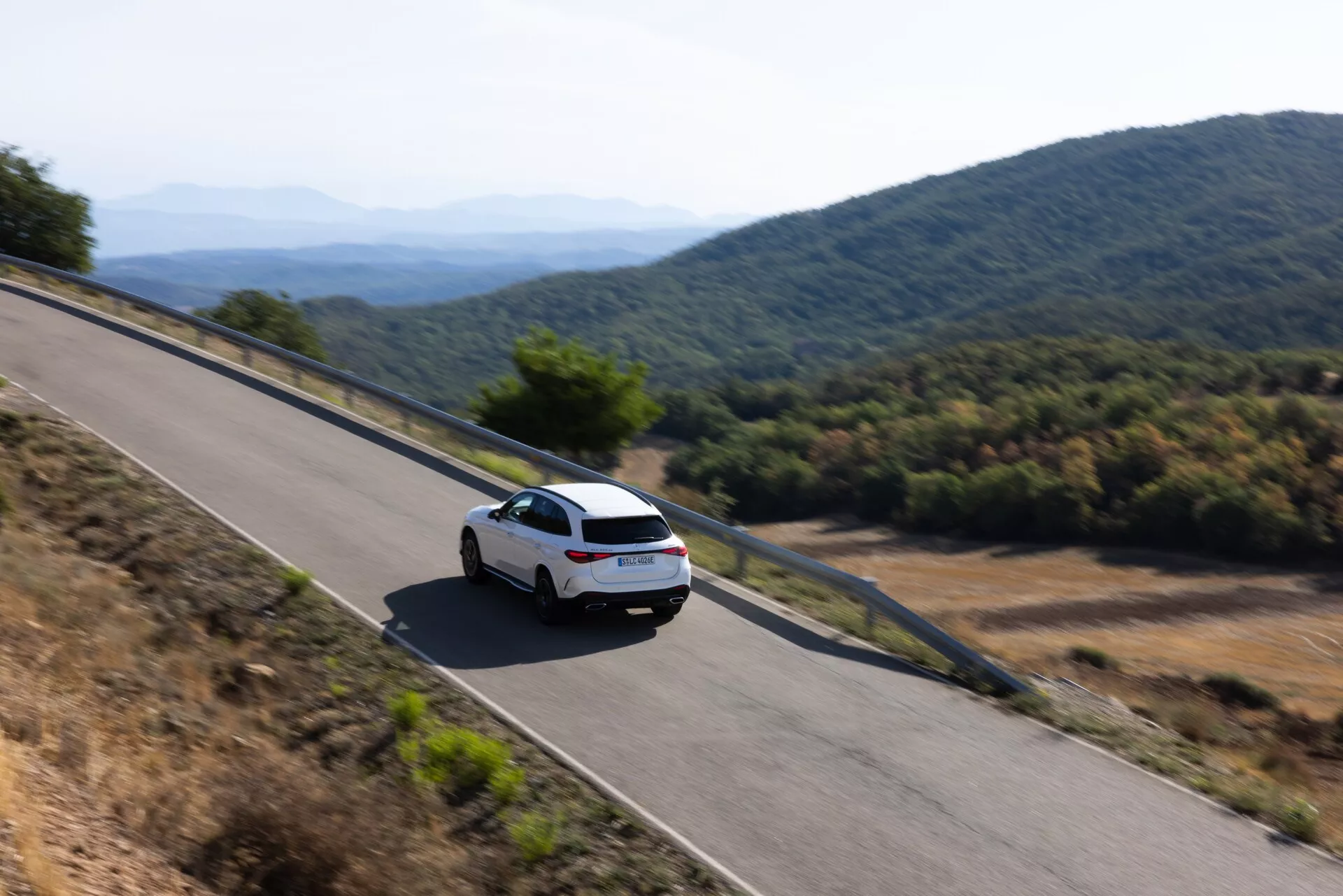 Högvinkel bakre trekvartsvy av en modern vit Mercedes GLC 2022 (254) som kör på en bergsväg. Bilen, som visas med en klar bakruta, baklyktor och en del av bagageutrymmet, fångar en känsla av rörelse med en lätt suddighet, omgiven av ett naturligt och varmt ljus under antingen morgon eller sen eftermiddag.