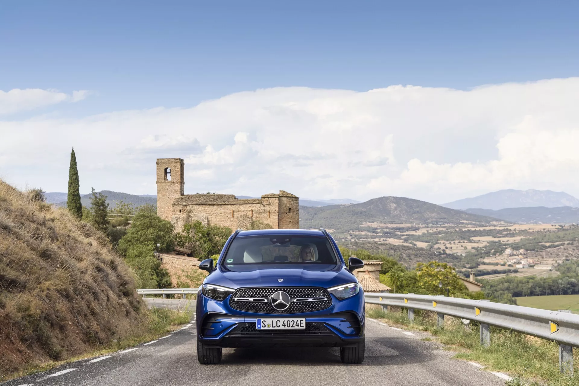 Mercedes-Benz GLC 254 från 2022 sedd från en vinkel med den främre och sidovyn synlig. Bilen har eleganta LED-strålkastare, en framträdande grill och sportiga lättmetallfälgar. Parkerad på en stadsgata med moderna byggnader i bakgrunden, under starkt dagsljus som framhäver dess metalliskt silverfinish.
