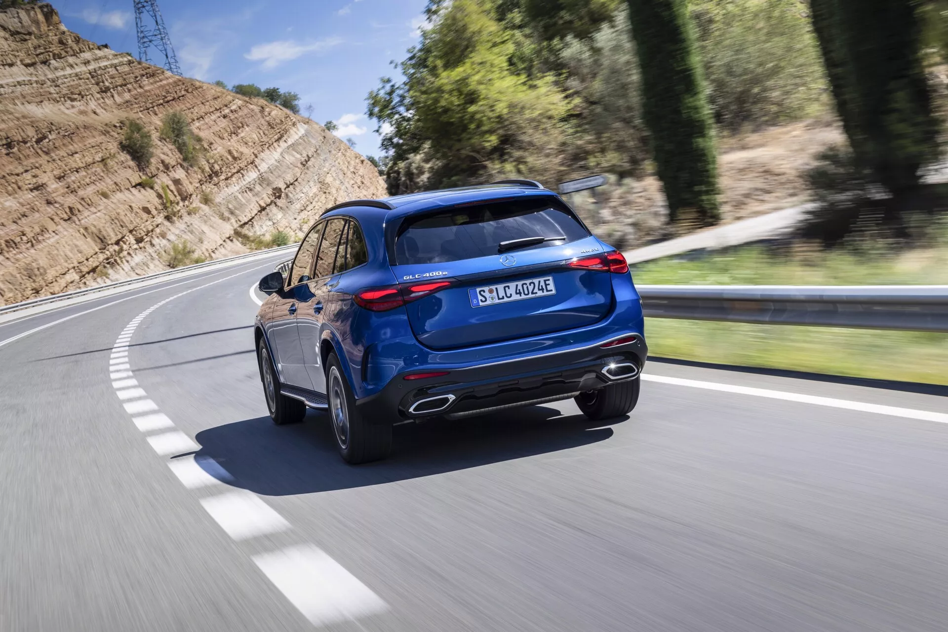 Bilden visar en dynamisk bakre trekvartsvy av en blå Mercedes GLC 2022 SUV som kör på en asfalterad väg som svänger åt vänster. Tagen från en något upphöjd vinkel visar fordonets höjdpunkter kromade avgasrör, en rejäl svartfodrad bakre stötfångare, eleganta LED-bakljus och en brant lutande bakruta. Den sportiga takspoilern, panoramataket och de synliga takräckena ger en högklassig attraktionskraft. Sidoprofilen avslöjar flerfälgslegeringshjul och lågprofildäck, vilket betonar vägprestanda. En laddningsport före bakljuset och EQ-märket indikerar plug-in hybrid eller elektriska kapaciteter, medan 4MATIC-märket anger fyrhjulsdrift. Den suddiga bakgrunden på grund av rörelse antyder att fordonet är i rörelse, vilket ger en känsla av hastighet och rörelse.