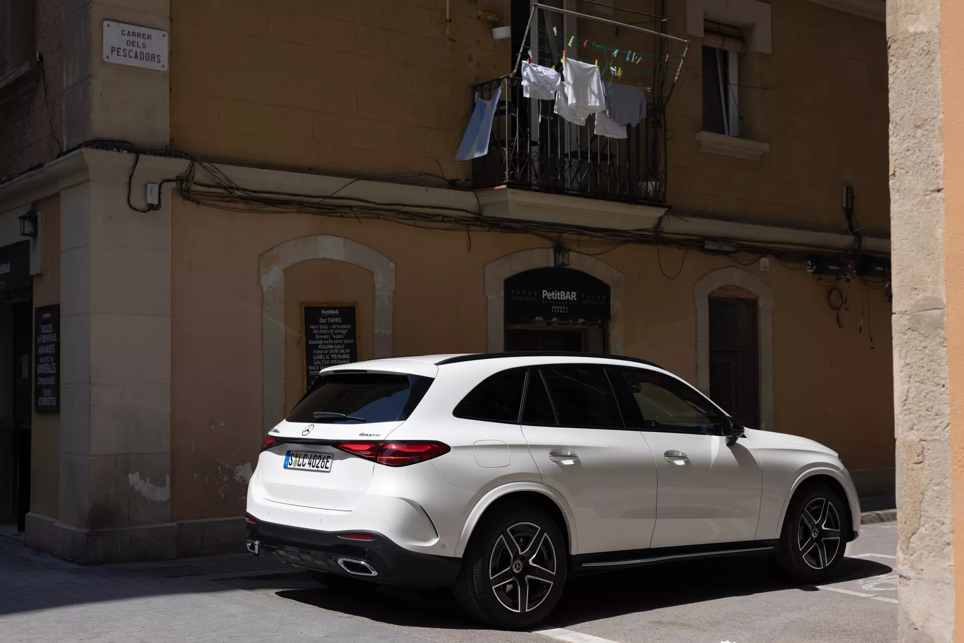 En samtida lyx-SUV parkerad på en stadsgata, fångad från en trekvarts bakvy, som visar upp eleganta LED-bakljus, dubbla avgasrör och en baklucka. Sidoprofilen lyfter fram stora distinkta fälgar, sportiga hjul, omdesignade sidospeglar med potentiella blinkers, ett soltak och ren enfärgad lack, vilket betonar moderna och prestandafunktioner i en urban miljö.