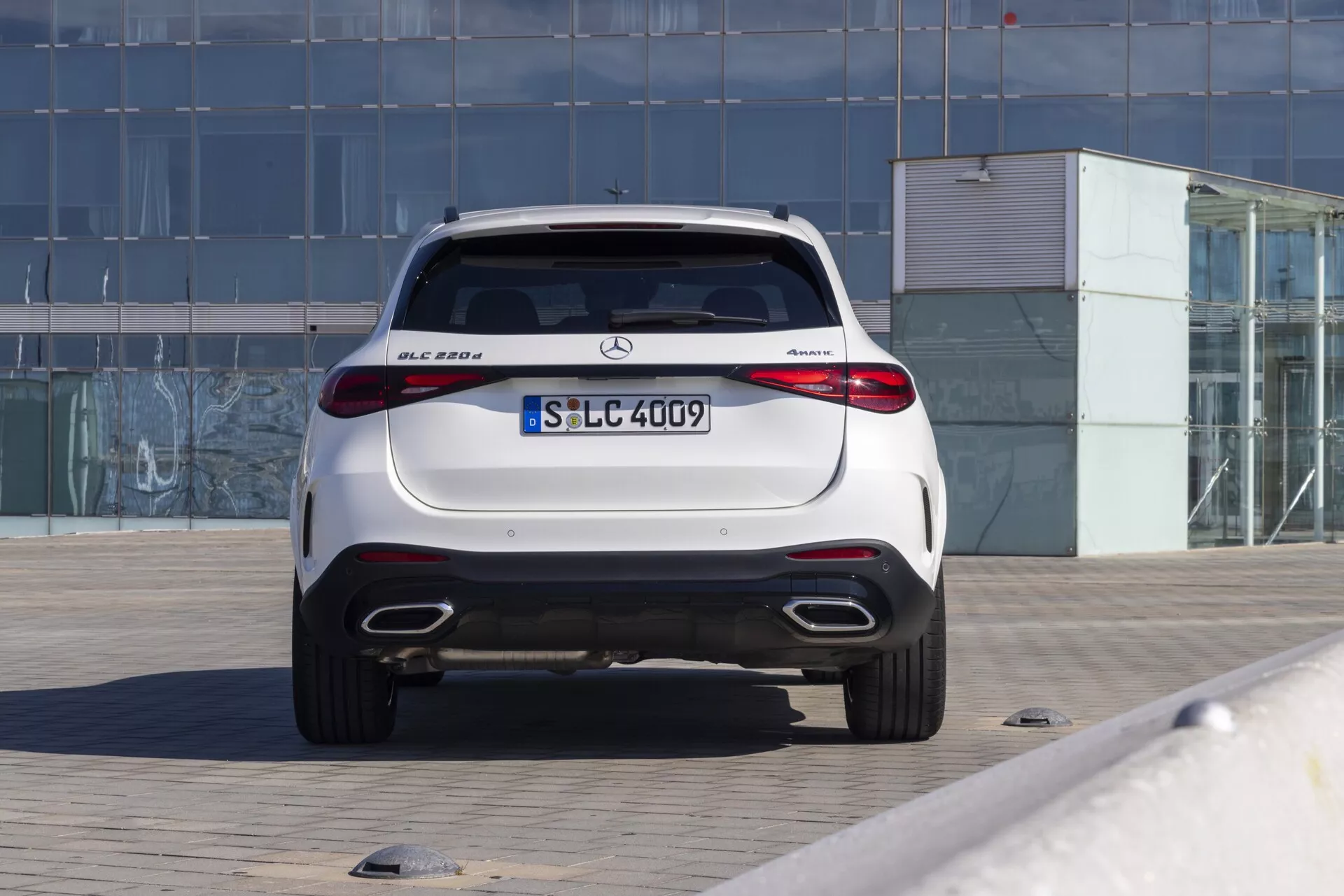 Bakre vy av en lyxig kompakt SUV Mercedes-Benz GLC 300 från 2022 med fyrhjulsdrift 4MATIC, parkerad på ett belagt område. Fotot visar fordonets aerodynamiska baklucka, integrerade bakspoiler, LED-bakljus med horisontell design, dubbla avgasrör, inbyggda parkeringssensorer, bakre diffusor och högblank finish. Bilen är centrerad i bildramen mot en bakgrund som visar en byggnad med glasfasad, vilket tyder på en urban eller kommersiell miljö. Den väl upplysta bilden framhäver det eleganta och sportiga designutseendet som är typiskt för moderna Mercedes-Benz-fordon.