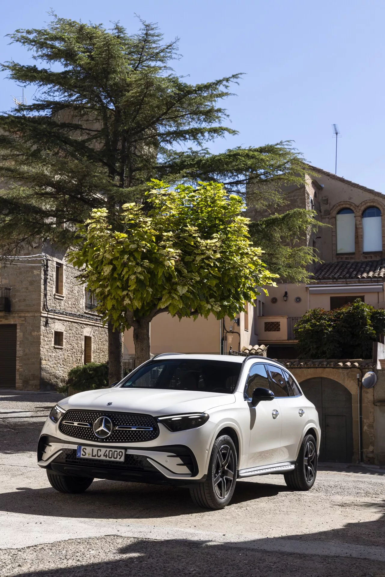 Vit Mercedes-Benz GLC 2022 (254) SUV parkerad på en kullerstensgata i en historisk europeisk stad, fångad från en frontal trekvartsvinkel. Synliga egenskaper inkluderar en stor Mercedes-Benz-emblem på grillen, LED-strålkastare, lättmetallfälgar, potentiella kameror/sensorer på den främre stötfångaren, kurvig karossdesign och speglar som möjligtvis rymmer ytterligare kameror eller svängsignaler. Miljön och det naturliga ljuset framhäver fordonets lyxiga utseende.