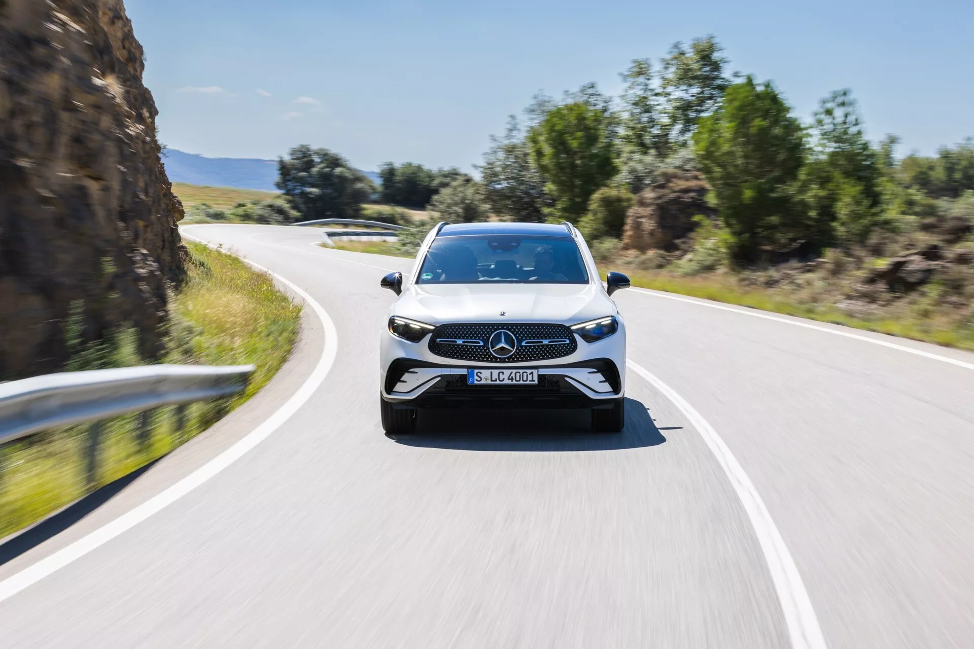 Frontal vy av en rörlig Mercedes-Benz GLC 2022, fångad något utanför centrum, som visar fronten och en del av sidorna. Bilden framhäver emblem och den eleganta grilldesignen, LED-strålkastare, stora luftintag och skulpterade linjer på motorhuven, vilket antyder en sportig och modern design. Den suddiga bakgrunden tyder på en känsla av fart, med bilen som färdas på en tvåfilig väg under en klar himmel, vilket framhäver dess dynamiska och avancerade tekniska egenskaper.