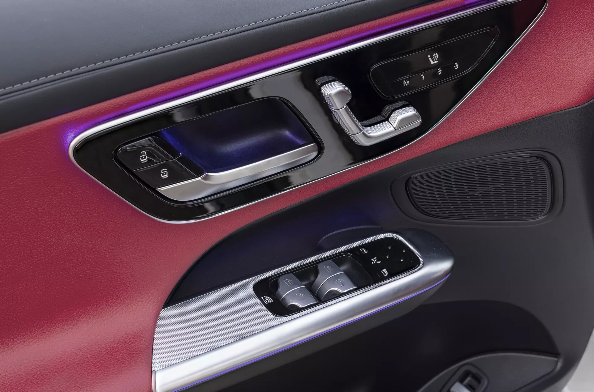 Närbild av förardörrpanelen på 2022 Mercedes GLC (254), som visar en blandning av material inklusive metalltrim och matta svarta ytor. Utrustad med färgad ambientbelysning, ergonomiskt utformad dörrhandtag i glansigt svart och metall, bakgrundsbelysta elektriska fönsterkontroller, eljusterbara sidospeglar och ett högkvalitativt högtalargaller integrerat längst ner, vilket betonar bilens lyxiga och teknologiskt avancerade interiördesign.