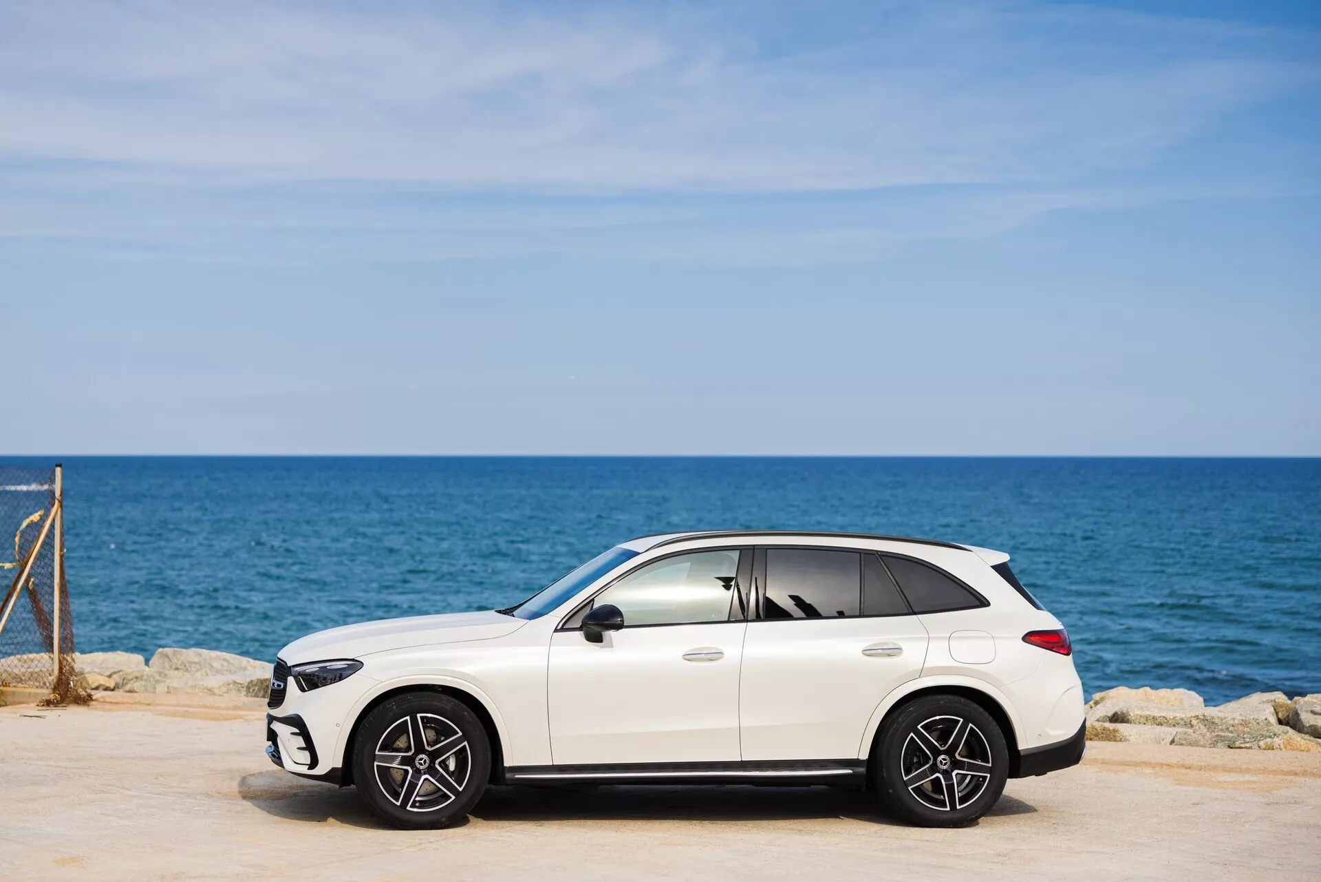 Bilden visar en vit, lyxig Mercedes GLC 2022 (254) crossover SUV som står parkerad på en asfalterad yta. Sidovy perspektiv framhäver passagerarsidans profil med synliga fram- och bakhjul av multi-ekrad design. I bakgrunden syns havet och en klarblå himmel. SUV:ens eleganta design innefattar sidospeglar, dörrhandtag och möjligen ett takfönster. Skuggor under bilen indikerar soliga förhållanden. Lätt tonade rutor bidrar till en lyxigare framtoning.