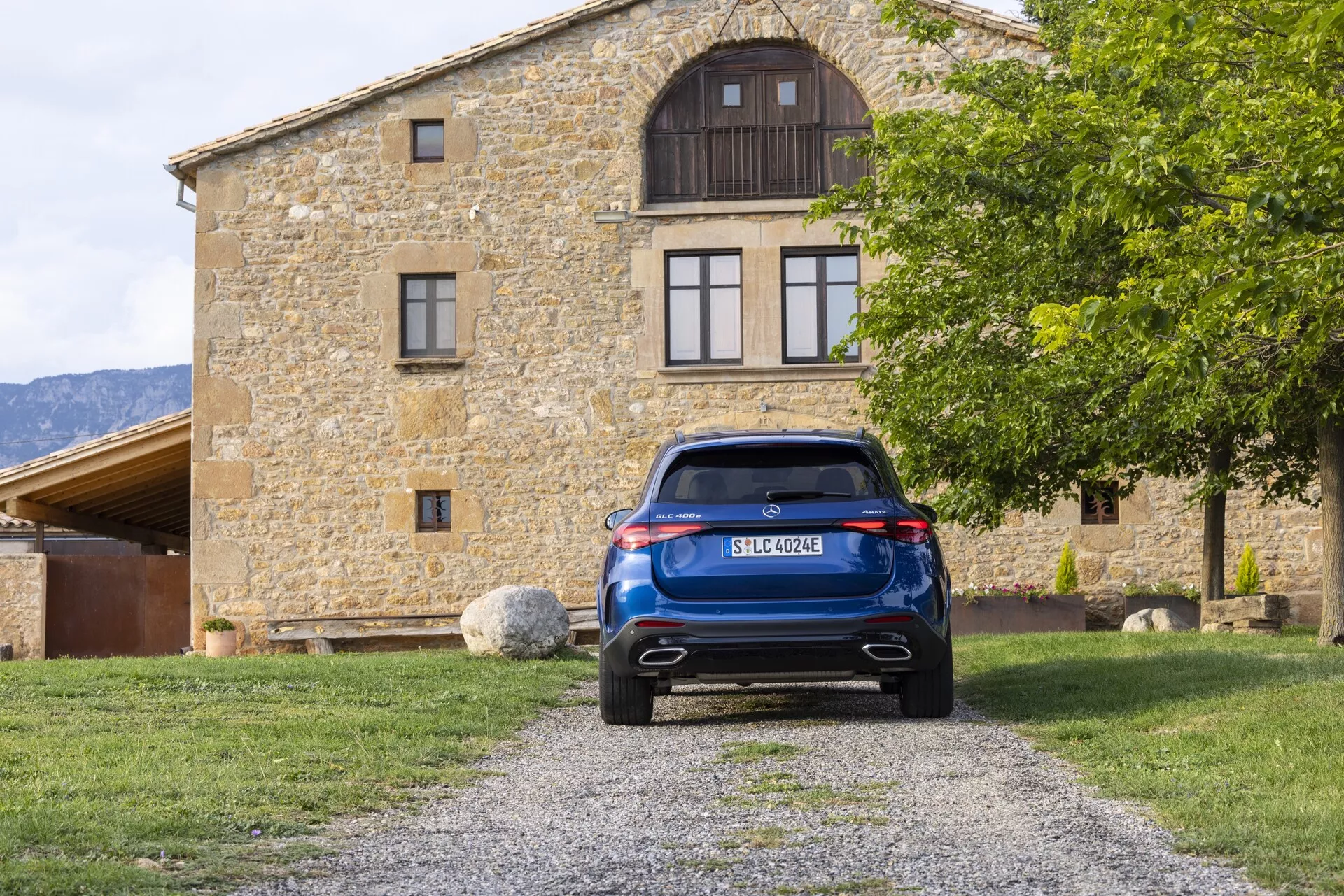 Bakifrånvy av en blå Mercedes GLC 2022 (254) parkerad på en grusväg, centrerad i kompositionen med en byggnad i bakgrunden. Bilden framhäver fordonets baklucka, bakre vindruta med torkare, eleganta LED-bakljus, bakre stötfångare med parkeringssensorer eller reflexer och dubbelt avgasrör. Funktioner inkluderar en högmonterad bromsljus, centralt placerad registreringsskylt, diffusorelement och takspoiler, som visar modern fordonsteknik och design.