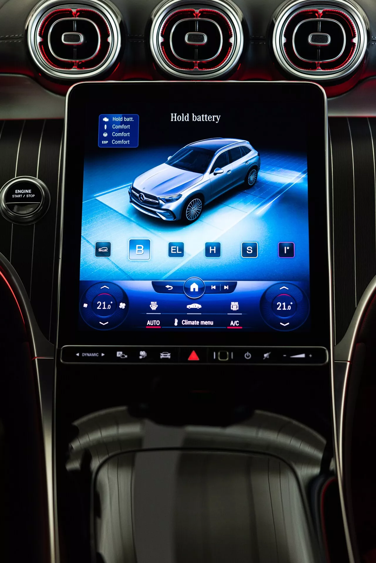 Fotografi av interiören i en Mercedes GLC 254 från 2022, med fokus på en centralt placerad, rektangulär vertikalt orienterad infotainmentdisplay på instrumentbrädan. Skärmen, omgiven av en blank svart ram, visar ett blått gränssnitt som möjligen indikerar hybrid- eller elfunktioner, med 'Hold battery' funktionen synlig. Gränssnittet inkluderar pekskontroller för klimathantering, medieuppspelning och bilinställningar, med knappar märkta 'B', 'E', 'H' och 'S' för olika körlägen eller batteriinställningar. Ovanför skärmen finns tre cirkulära luftventiler med röda accenter, vilket indikerar en premiumdesign. Den nedre delen av skärmen har tvåzons klimathantering inställd på 21°C, med ikoner för autoläge, luftcirkulation och luftkonditionering som ger ett modernt, högklassigt estetiskt intryck. Scenen visar också en smal silverram och en del av växelspaken tillsammans med en glimt av en mugghållare eller förvaringsfack.