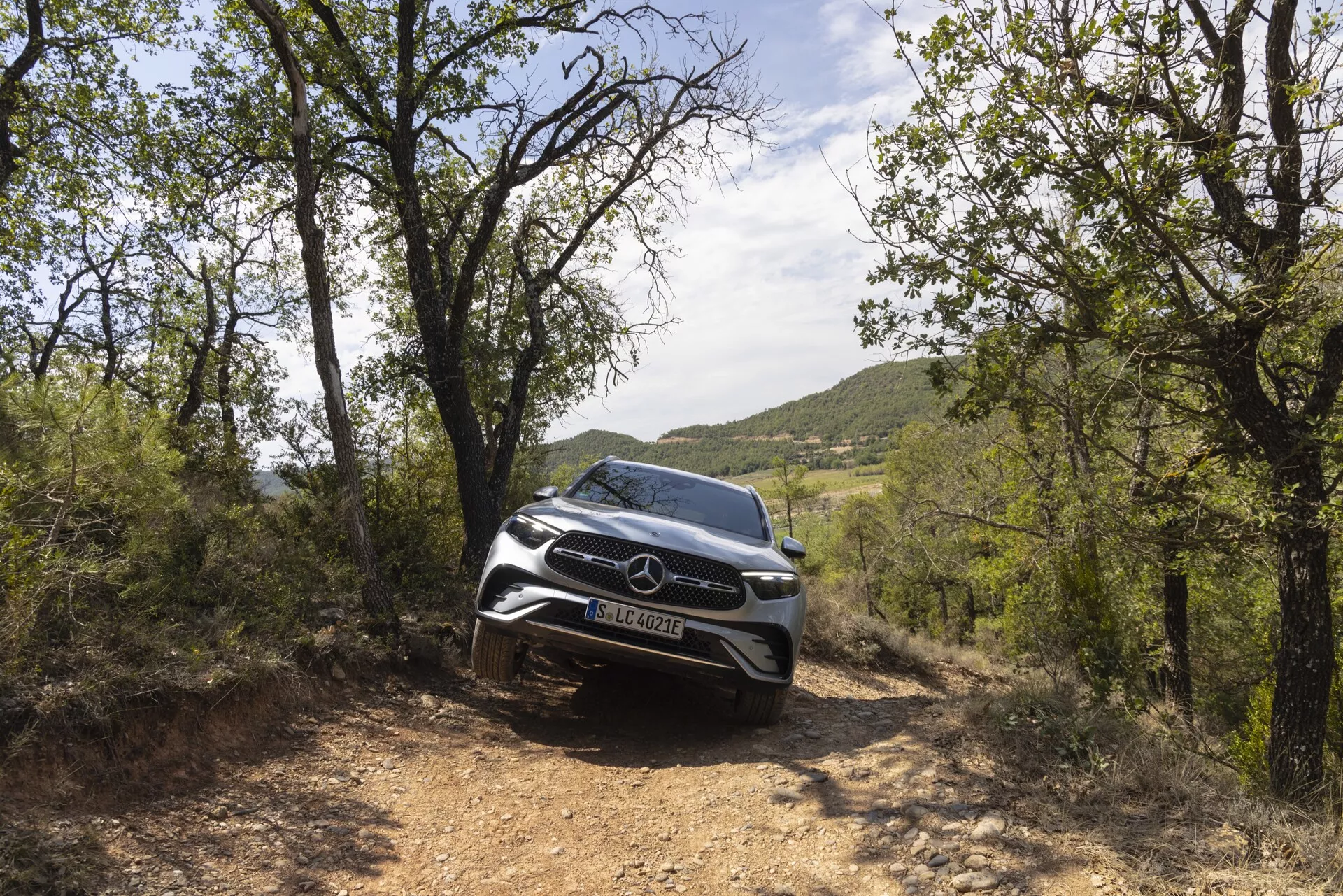 Färgbild av en Mercedes-Benz GLC 2022 SUV i en naturlig miljö, tagen från en låg framvinkel som betonar dess robusta utseende. Fotot fokuserar på bilens främre fasad, grill och moderna LED-strålkastare. Fordonet är på en grusväg omgiven av lövverk, vilket framhäver dess terrängkapacitet och eleganta design. Främre sensorer och kameror antyder avancerade säkerhetsfunktioner.