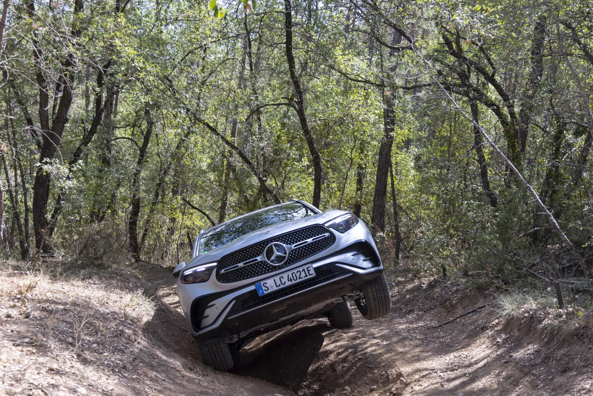 Mercedes-Benz GLC SUV 2022 navigerar en utmanande, skogsklädd terrängstig från en trekvarts framvinkel, visar full grill, emblem, strålkastare, del av huven och främre stötdämpare med slitskydd; passagerarsidan mer synlig med lutande hållning på ojämn mark, omgiven av träd och buskar som fångar SUV:ns terrängförmåga.