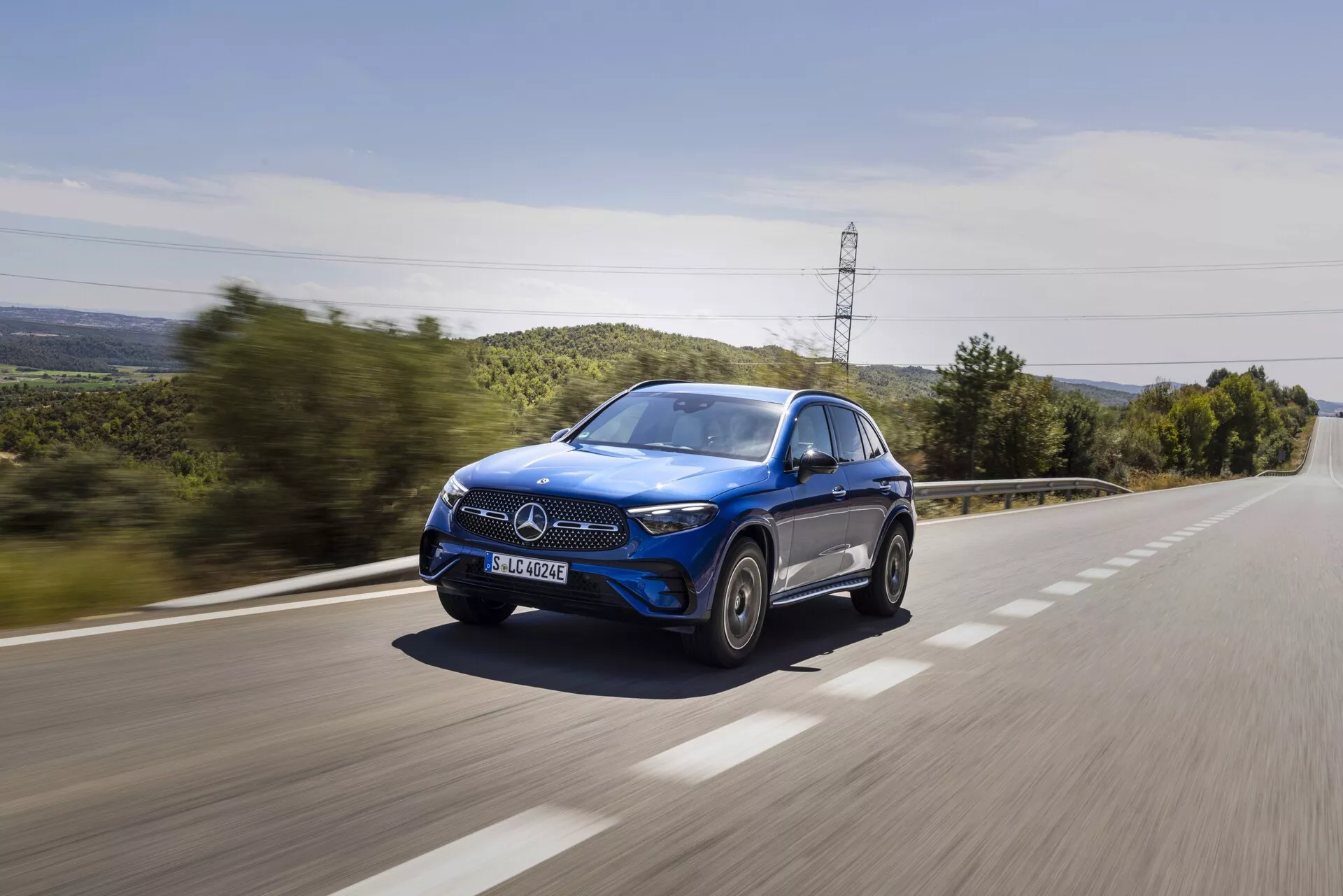 En blå Mercedes-Benz GLC 2022 SUV som kör med hög hastighet på en landsväg, fångad från en trekvarts framvinkel, visar frontgrillen, motorhuven, LED-strålkastarna, sidospegeln på förarsidan, hjulen och fönstren, med en rörelse-sudd-effekt på vägbanan och grönskan i bakgrunden, vilket framhäver bilens dynamiska och lyxiga design.