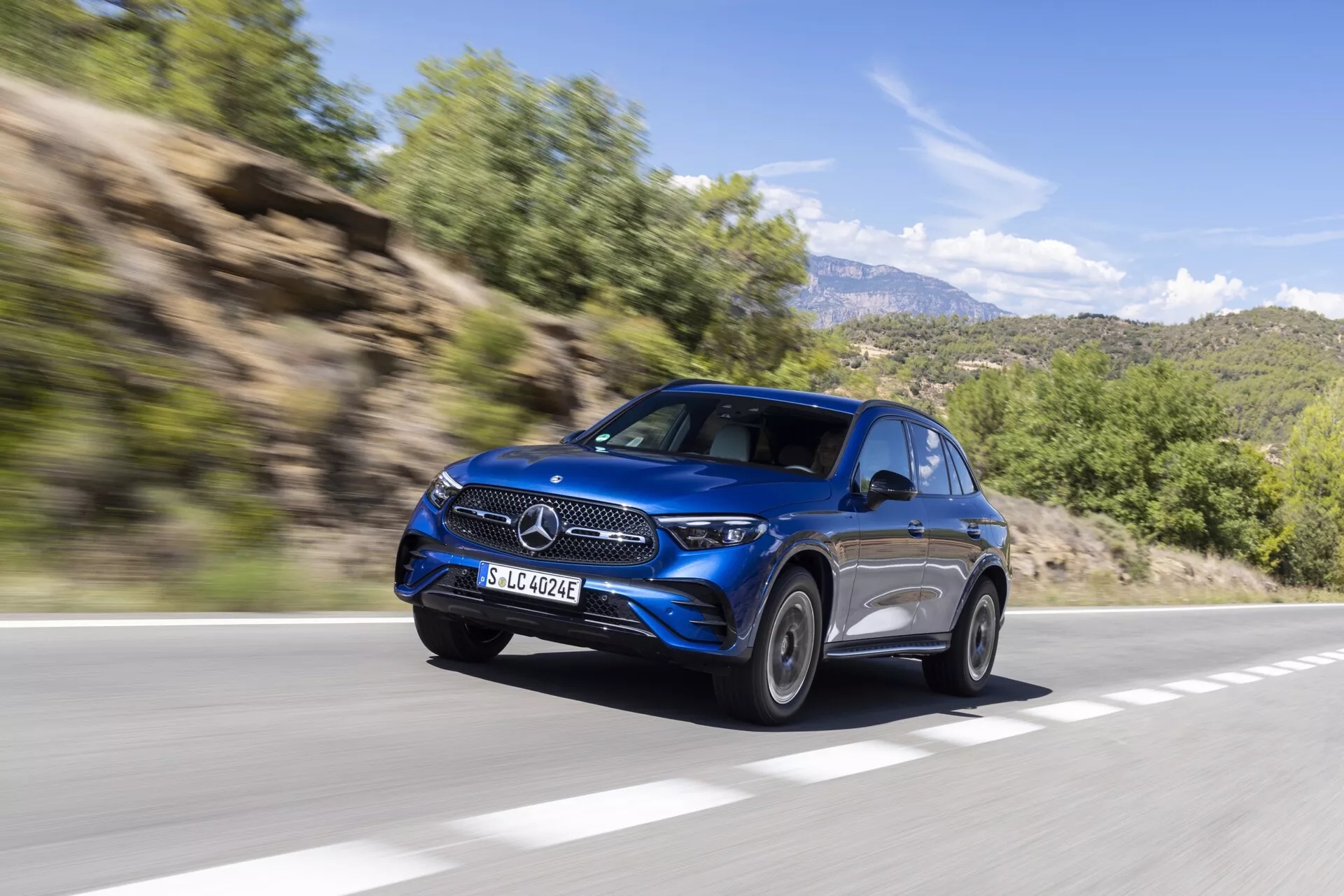 Dynamisk bild av en blå Mercedes GLC 2022 (254) SUV i rörelse, sedd från vänster framsida. Bilden visar dess eleganta strålkastare, framträdande grill med centralt emblem, lyxiga lättmetallfälgar, aerodynamiska karosslinjer och avancerade sidospeglar. Fordonet kör på en asfalterad väg i ett lantligt bergsområde och rörelseoskärpan i bakgrunden med lövverk och klippiga kullar bidrar till ett intryck av hastighet och modern lyx.