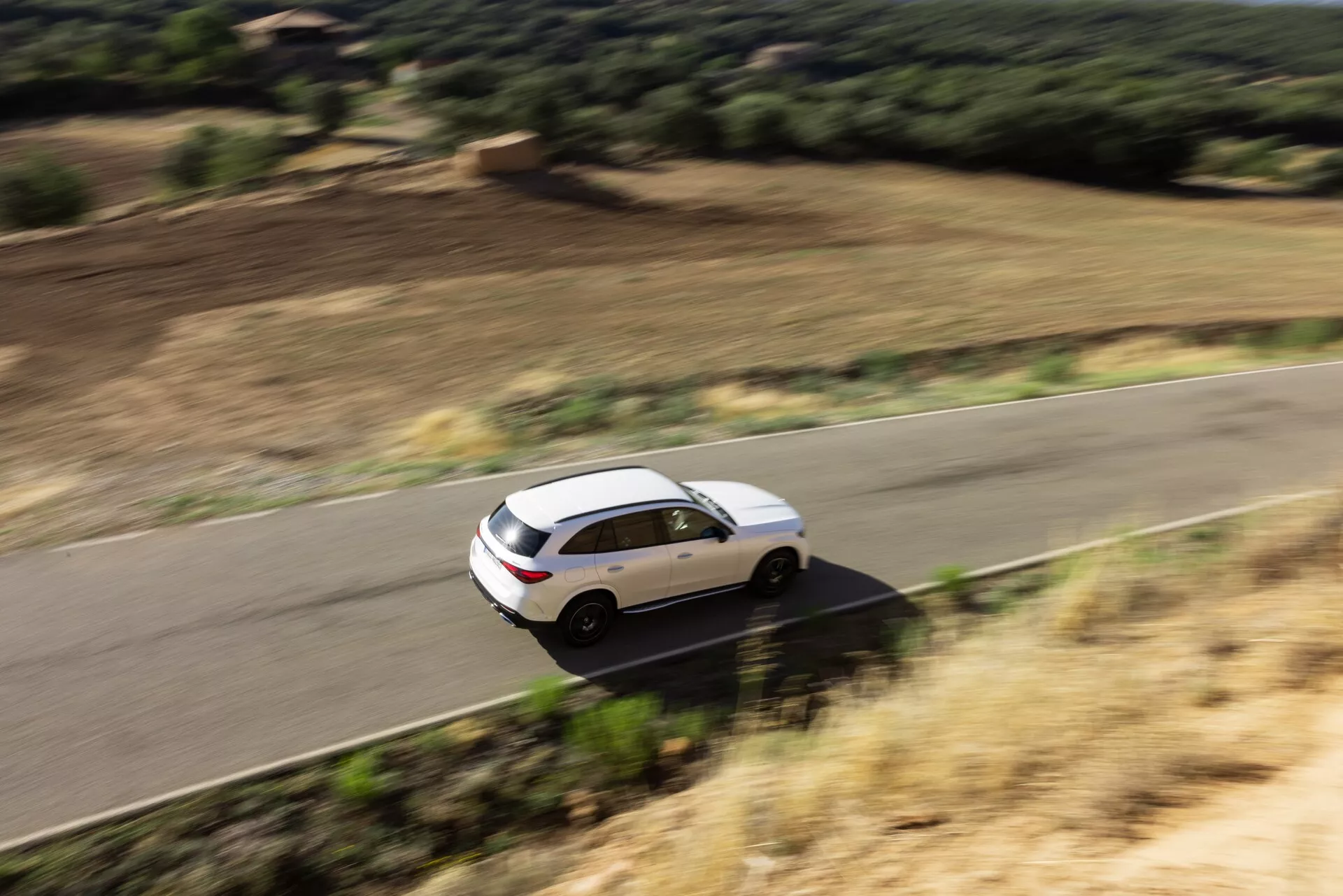 Hög vinkelvy av en fyrdörrars vit Mercedes GLC 2022 SUV i rörelse på en slingrande landsväg omgiven av naturligt landskap. Fordonet har en strömlinjeformad aerodynamisk kaross, panoramasoltak, lättmetallfälgar, tonade bakrutor och sidospeglar med integrerade blinkers. Rörelseoskärpan antyder hastighet och dynamisk prestanda.