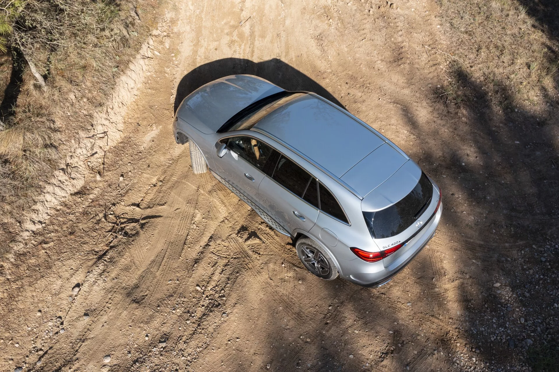 Övergripande vy av en silverfärgad Mercedes GLC 2022, som visar dess moderna design med stängt soltak, bakre antenn och femekrade aluminiumfälgar. Bilen, som är placerad på en dammig landsväg, lyfter fram dess möjliga hatchback- eller sportback-kaross, med skuggor som indikerar morgon- eller eftermiddagssolljus.