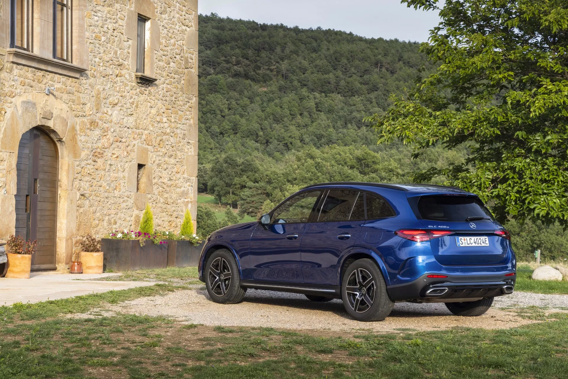 2022 Mercedes-Benz GLC lyxcrossover SUV på grusuppfart bredvid historisk stenbyggnad, från trekvarts bakperspektiv, visar djupblå lackering, LED-bakljus, dubbla avgasrör, lättmetallfälgar och sportig taklinje under middagssol.
