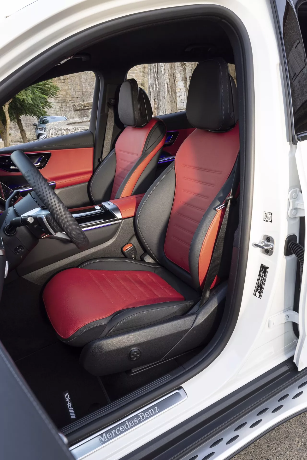 Interiörvy av 2022 Mercedes-Benz GLC, som framhäver de tvåfärgade sportiga framsätena med glänsande läder, avancerad styrpanel för sätena på förardörren som är öppen, samt det delvis synliga avancerade multifunktionella ratten och elegant instrumentbräda med anpassningsbar omgivningsbelysning.