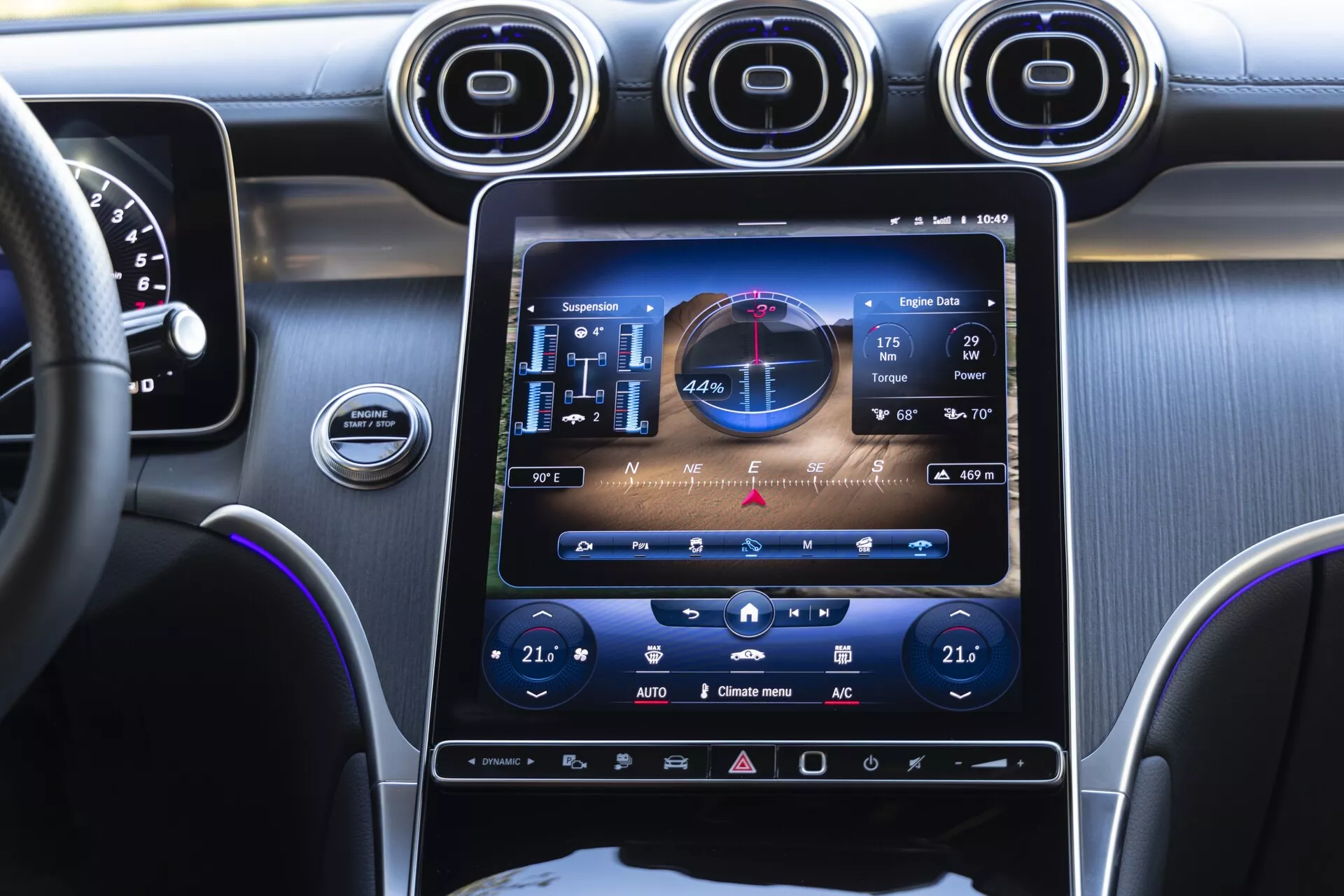 Ögonhöjdsvy från framsätet på passagerarsidan i en Mercedes GLC 2022-interiör, som framhäver den centrala konsolen med ett stort vertikalt pekskärminfoystem som visar GPS, ljud och klimatkontroller. Tre runda metalliska luftventiler ovanför skärmen, antydan till instrumentpanel och ratt till vänster, med lila stämningsbelysning som betonar konsolen och instrumentpanelen.