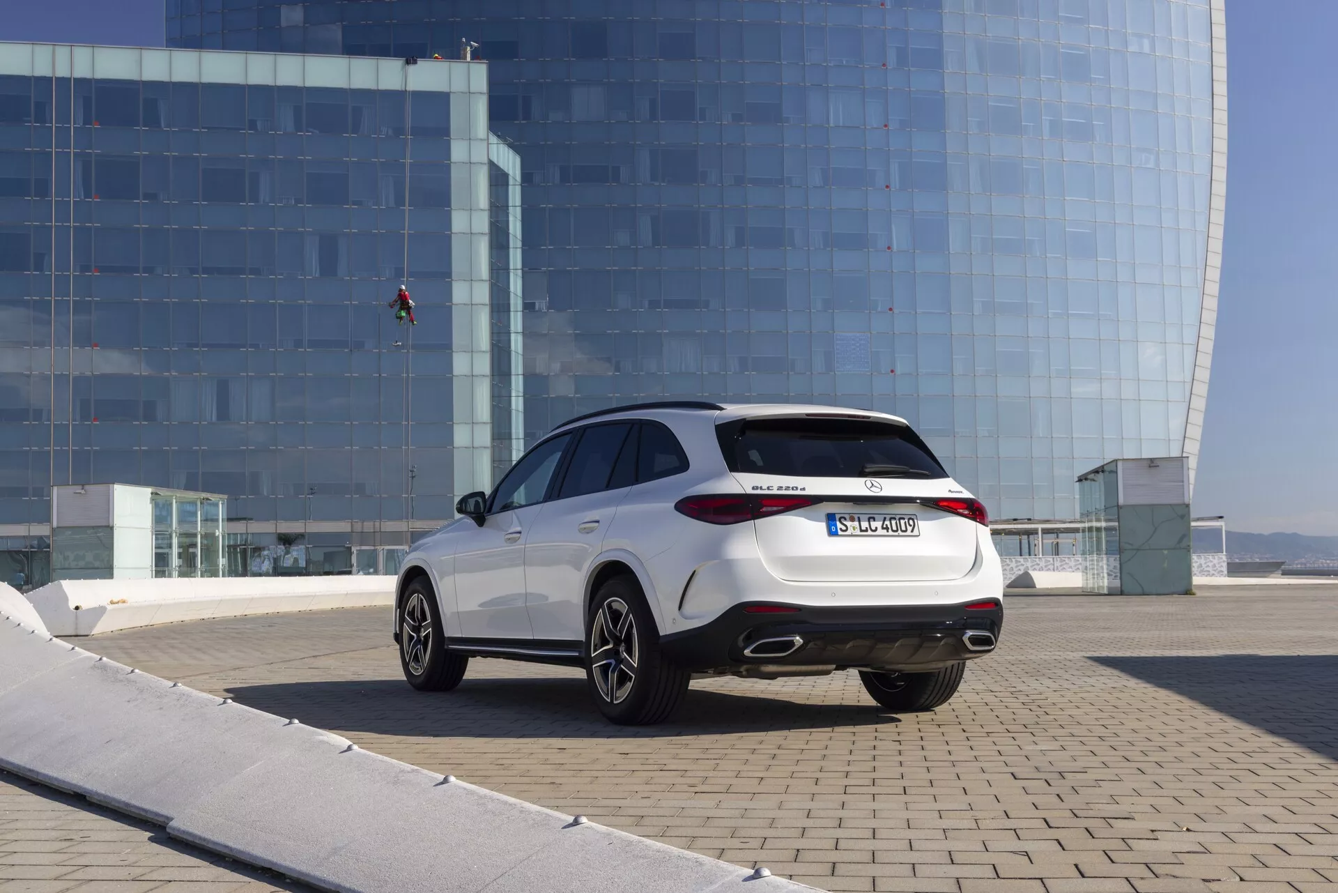 På bilden syns en modern Mercedes GLC 2022 (254) bil sedd från en utomhus trekvart bakvy, diagonalt placerad med bakdelen närmare kameran, vilket visar upp moderna LED-bakljus, ett dubbelt avgasrör, lätt tonade bakrutor, en jämnt sluttande taklinje, flereker lättmetallfälgar med lågprofildäck, allt i en urban miljö med en reflekterande glasbyggnad i bakgrunden.