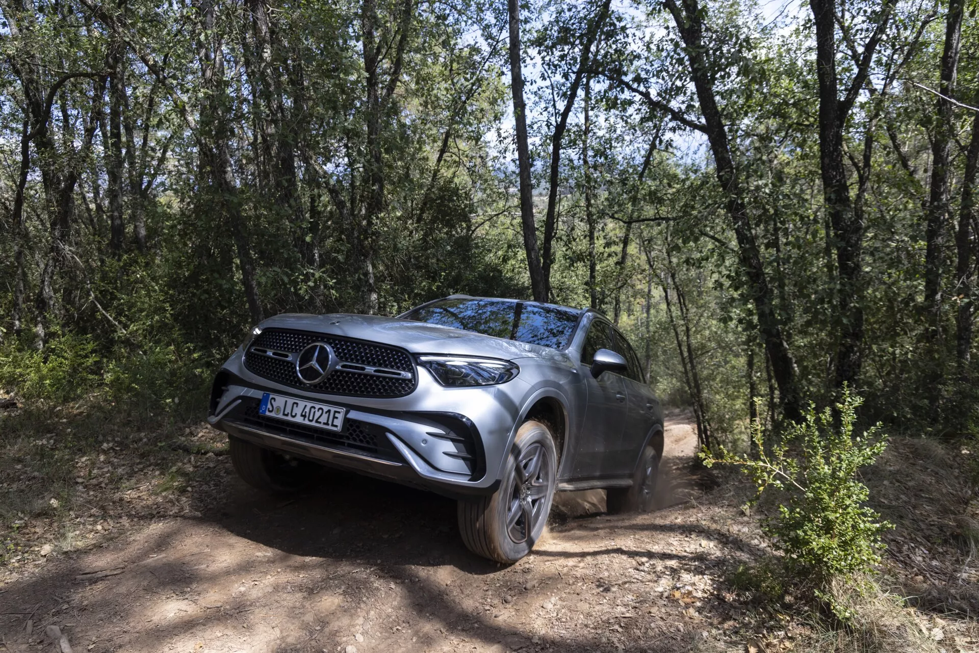2022 Mercedes GLC SUV positionerad på en grusväg omgiven av ett skogsområde, fångad från en fram-trekvarts vinkel som betonar dess framträdande frontgrill med varumärkesemblem, LED-strålkastare, lättmetallfälgar och hög markfrigång, vilket framhäver dess terrängegenskaper i en naturlig miljö av träd och torr terräng.