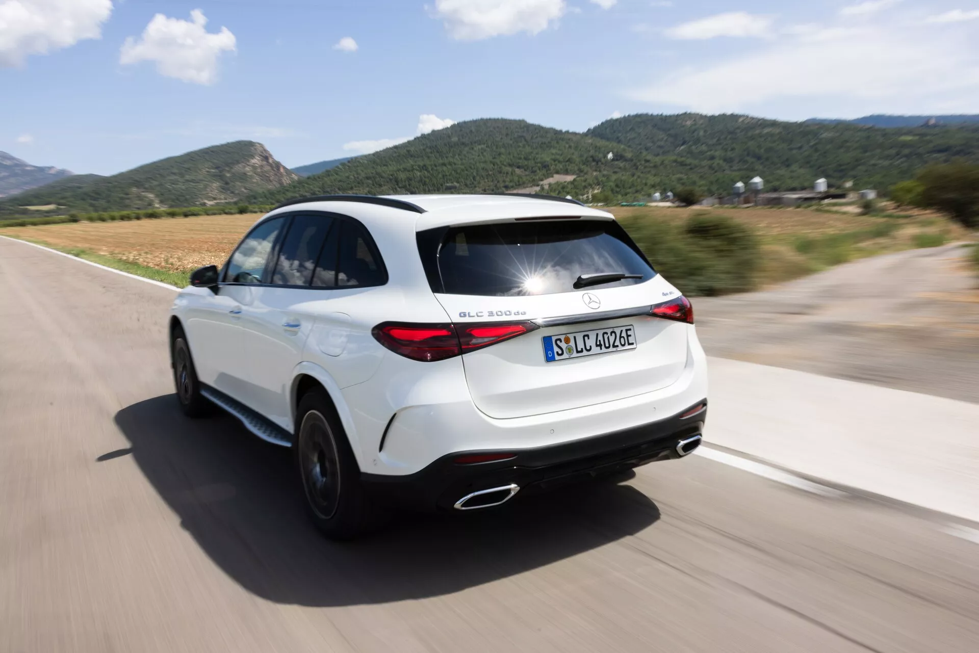 Dynamisk bakre trekvarts vy av en vit SUV, Mercedes GLC 254 från 2022, i rörelse med panoreringseffekt. Kameravinkeln fångar elegant design och moderna baklyktor, visar höger bakre kvartsdel, baklyktor, bakre hjul och del av höger sidoprofil på en välasfalterad väg. Betonar den elektriska/hybrida varianten med 'EQ'-märkning, sportig hållning, aerodynamiska sidokjolar, dubbla avgasrör och coupe-liknande taklinje. Osuddad bakgrund och hjulen framhäver bilens hastighet och rörelse.