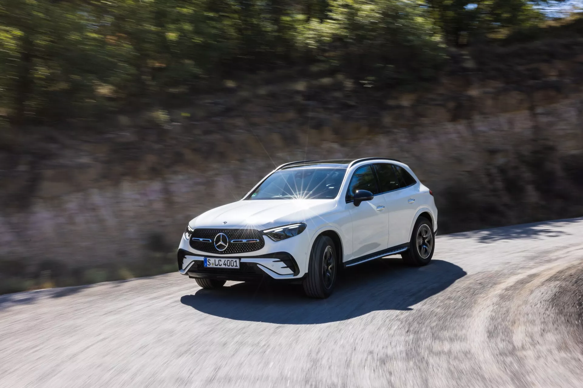 Dynamisk bild av en vit Mercedes-Benz GLC 2022 SUV i rörelse, fångad under dagsljus från en trekvart framvinkel. Framträdande detaljer inkluderar den främre grillen med ett framträdande emblem, distinkta LED-strålkastare, främre stötfångare med synliga luftintag, skulpterad motorhuv med upphöjda linjer och sidoprofilen upp till bakdörrarna. Hjulen är skildrade i rörelse med en lätt oskärpa, vilket antyder fart. Sidospeglarna är aerodynamiskt utformade. Miljön verkar suddig på grund av fordonets rörelse, vilket betonar bilens dynamiska och snabba karaktär.