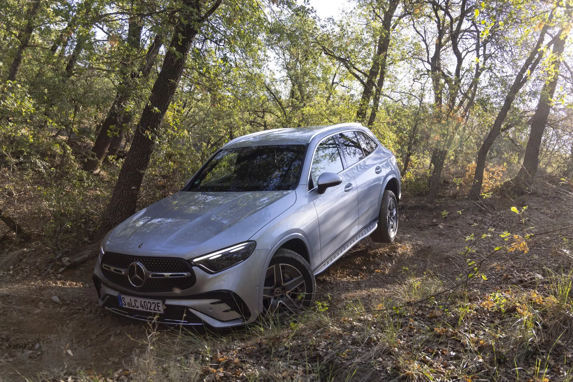 Bild av en silverfärgad 2022 Mercedes GLC SUV som är placerad utomhus i ett skogsområde och visar dess terrängegenskaper. Fordonet är vinklat åt vänster vilket ger en trekvarts vy av den främre och sidoprofilen, inklusive frontgallret, skarpa LED-strålkastare, konturerad främre stötfångare, motorhuv, legeringshjul med allterrängdäck och släta linjer. Naturligt solljus filtrerar genom träden och framhäver den moderna, aerodynamiska designen samt antyder avancerade körhjälpsfunktioner placerade bakom gallrets emblem.
