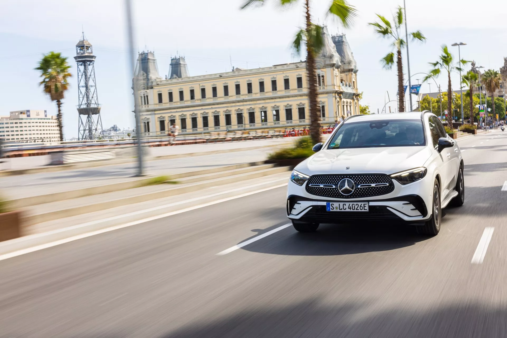 Mercedes-Benz GLC 2022 (254) lyxig elektrisk sedan i vitt med svart tak, fångad i rörelse i en dagtid stadsinställning med palmer och historiska byggnader i bakgrunden, med panoreringsteknik för att hålla fordonet skarpt mot den rörelseoskarpa bakgrunden. Med det karakteristiska Mercedes-Benz-emblemet på grillen, förarstödsensorer, tunna dagsljuslampor, eleganta LED-strålkastare och integrerade blinkers i speglarna.