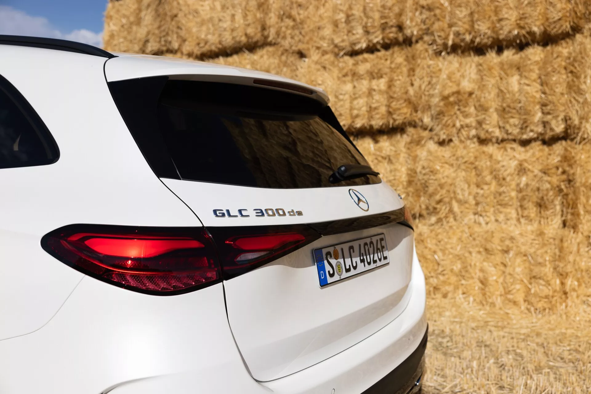 Närbild på en vit Mercedes-Benz GLC 300 d SUV sedd från trekvarts bakre vinkel, som visar högra sidan av bakdelen inklusive den bakre bagageluckan, den upplysta baklampan och en del av de bakre fönstren. Fordonet är parkerat i en lantlig miljö med höbalar i bakgrunden. Synliga funktioner inkluderar modellnamnet 'GLC 300 d', Mercedes-Benz-loggan och en europeiskt format registreringsskylt, som belyser fordonets eleganta designlinjer och moderna belysning.