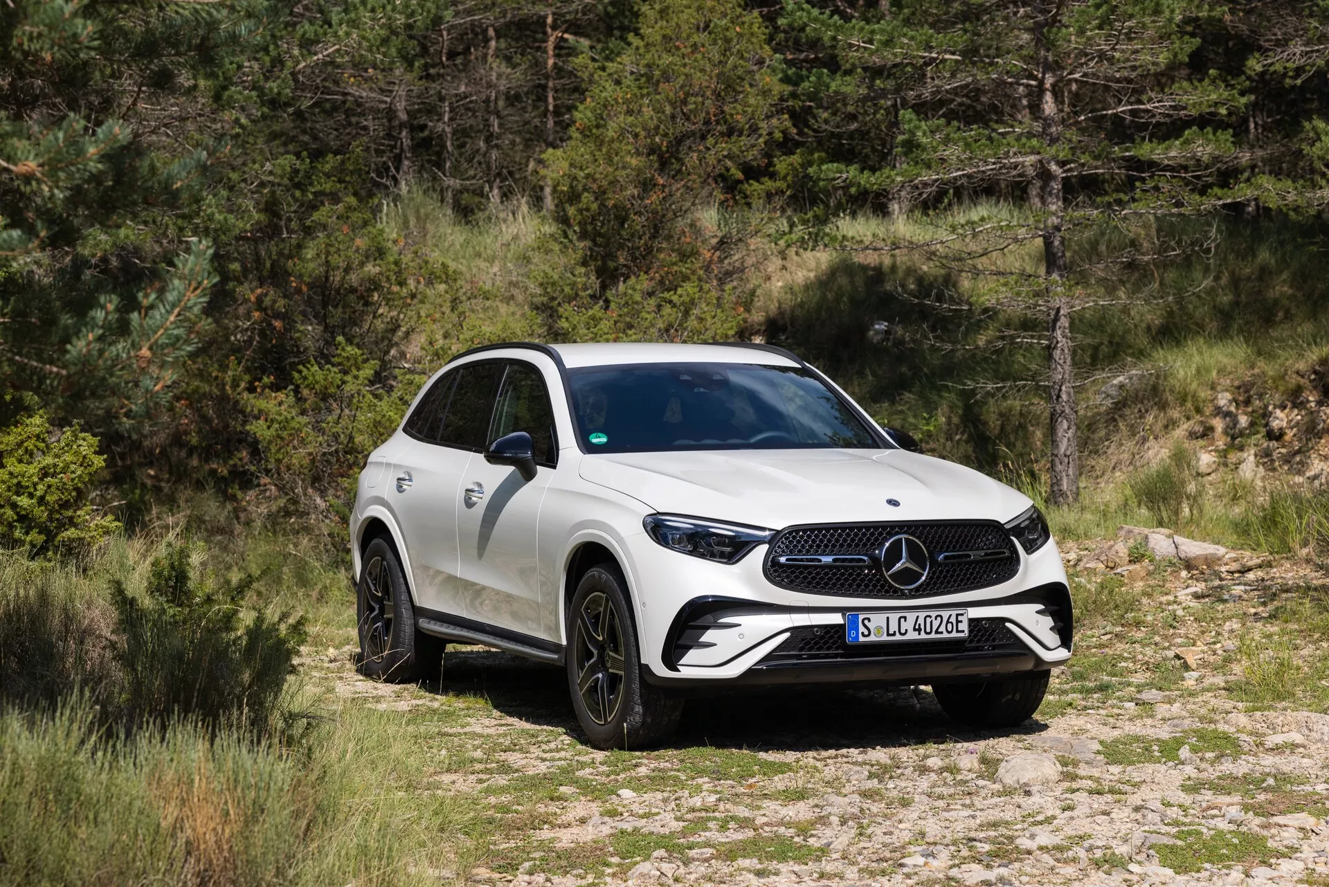 Bild av en Mercedes-Benz GLC SUV från 2022 i en naturlig miljö, placerad på en grus- eller jordväg med vegetation i bakgrunden. Fordonet är fotograferat i en trekvarts framvy, som visar frontgrillen med ett stort emblem, skarpa LED-strålkastare och en del av sidokarossen. SUV-en har flerspokes svarta legeringshjul, sidobackspeglar med integrerade blinkers och en uppåtgående midjelinje som leder till en sportig, avsmalnande bakdel. Karossen är i solid vit färg, och bilden antyder att denna modell är en del av Mercedes-Benz elektrifierade serie, vilket indikeras av 'EQ' -märket på framskärmen.