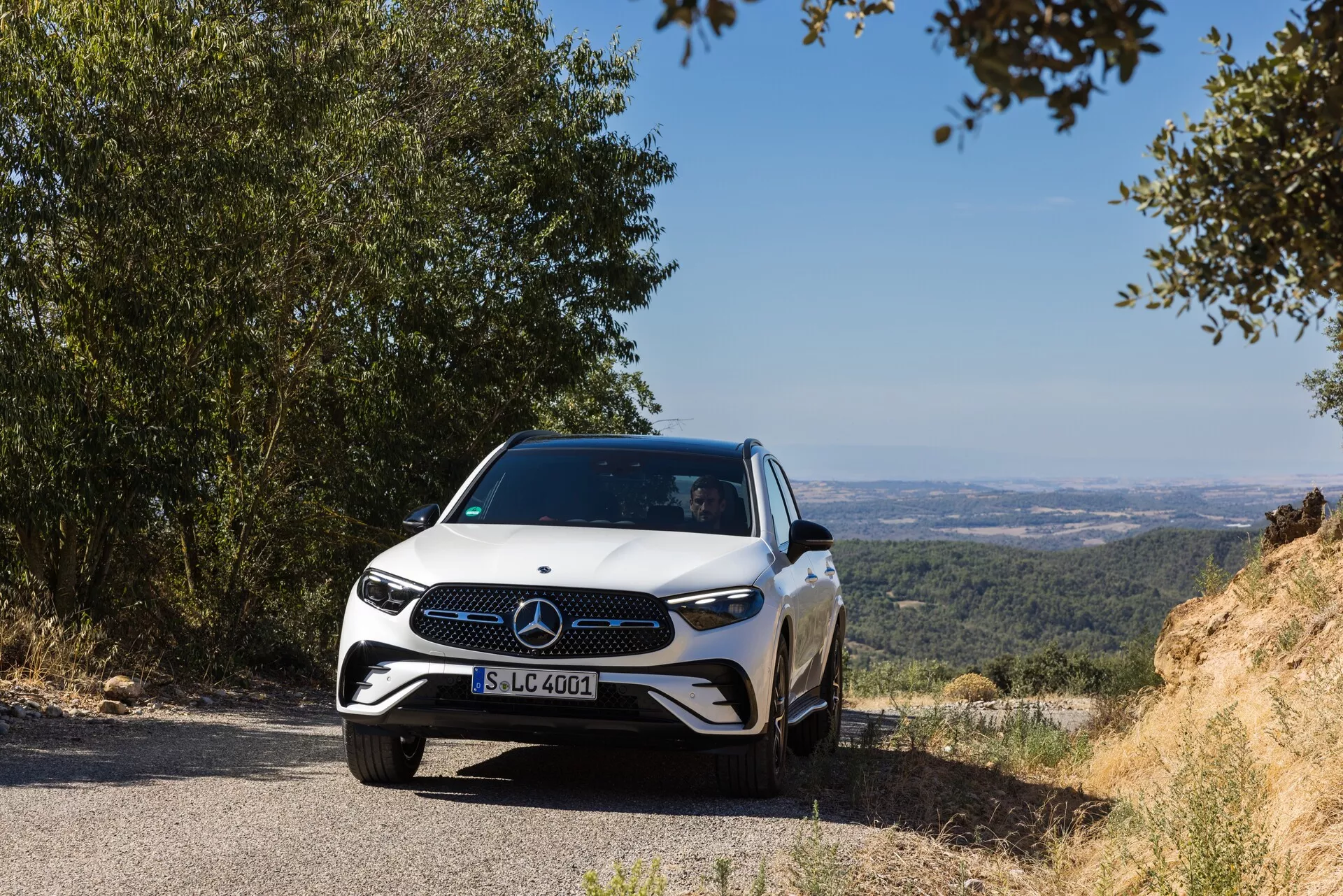 En modern Mercedes-Benz GLC 2022 är placerad på en smal väg med en naturlig, kuperad bakgrund som ger en vy över en dal eller slätter. Bilens främre emblem och grilldesign är tydligt synliga, fotograferade från en låg frontal vinkel något åt vänster, som framhäver frontgrillen, strålkastarna och en del av motorhuven. LED-strålkastare, en distinkt grill med ett stort emblem och en sportig stötfångardesign betonar bilens avancerade och eleganta egenskaper. Dagsljusfotot med naturlig belysning skapar en känsla av fridfullhet, och förmedlar avancerade förarassistanssystem antydda av emblemet. Den smala aerodynamiska profilen framhävs av motorhuvens linjer som smälter samman med de skulpterade sidorna, fotograferade på ett avstånd på cirka 3-5 meter från kameran.
