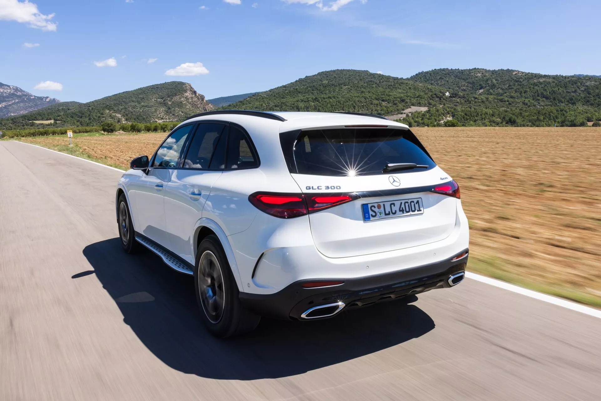 Hög vinkel i trekvarts bakifrån av en vit Mercedes-Benz GLC 300 SUV på väg. Bilden visar designelement som identifierbara märken, LED-bakljus, kromavgasrör, femekrade legeringshjul och en distinkt takspoiler. SUV:en är i rörelse på en solig dag, vilket framhäver dess dynamiska kapacitet och sportiga estetik.