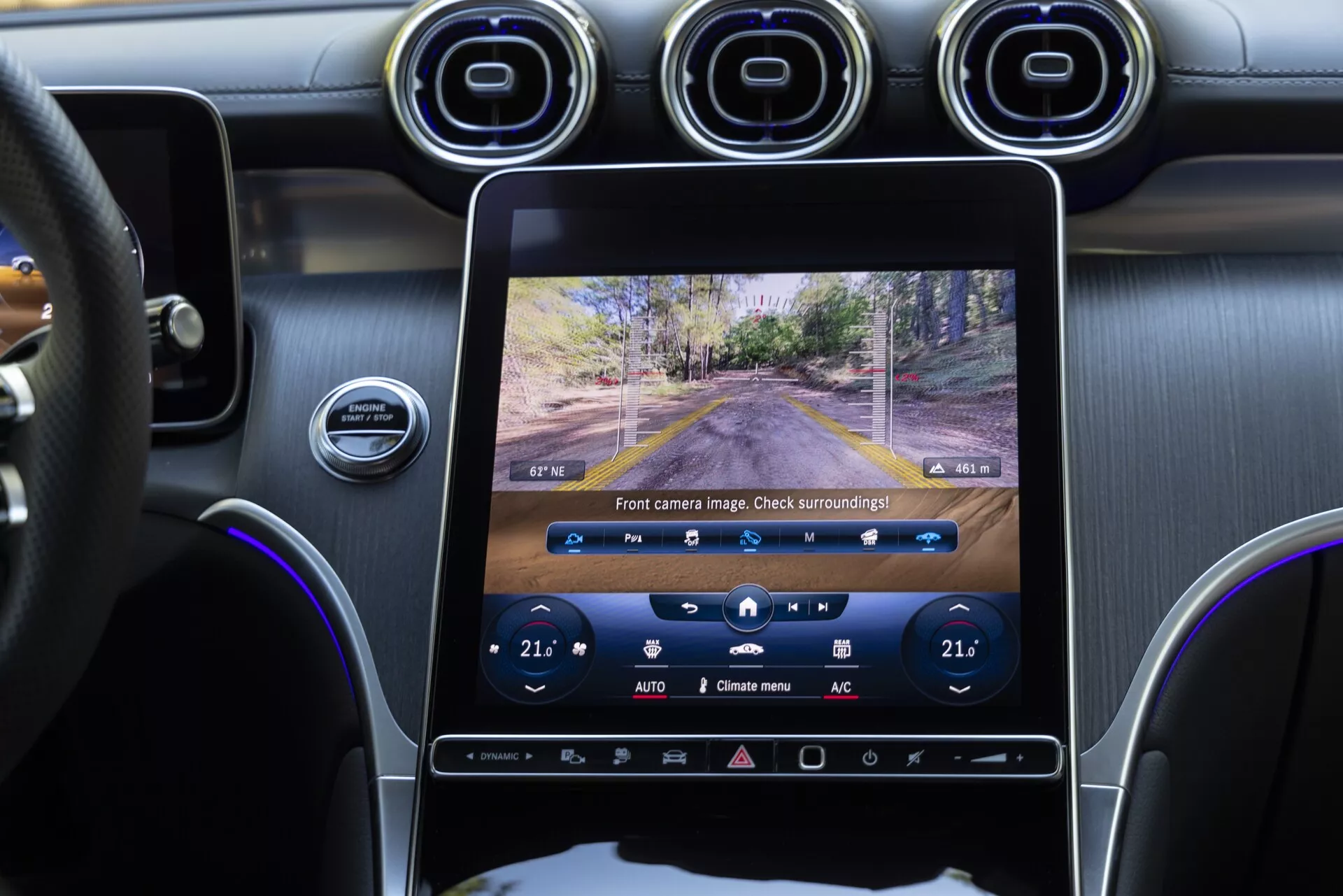 Interiörvy av 2022 Mercedes GLC som fokuserar på mittkonsolen och infotainmentsystemet, tagen från baksätets passagerarperspektiv. Den stora vertikala pekskärmen visar en live-feed från frontkameran, med klimatkontroller inställda på 21°C synliga nedanför. Instrumentbrädan, ratten och en del av förardörren är också synliga, som visar en kombination av eleganta digitala gränssnitt och fysiska knappar, cirkulära metallventiler och en läderklädd ratt.