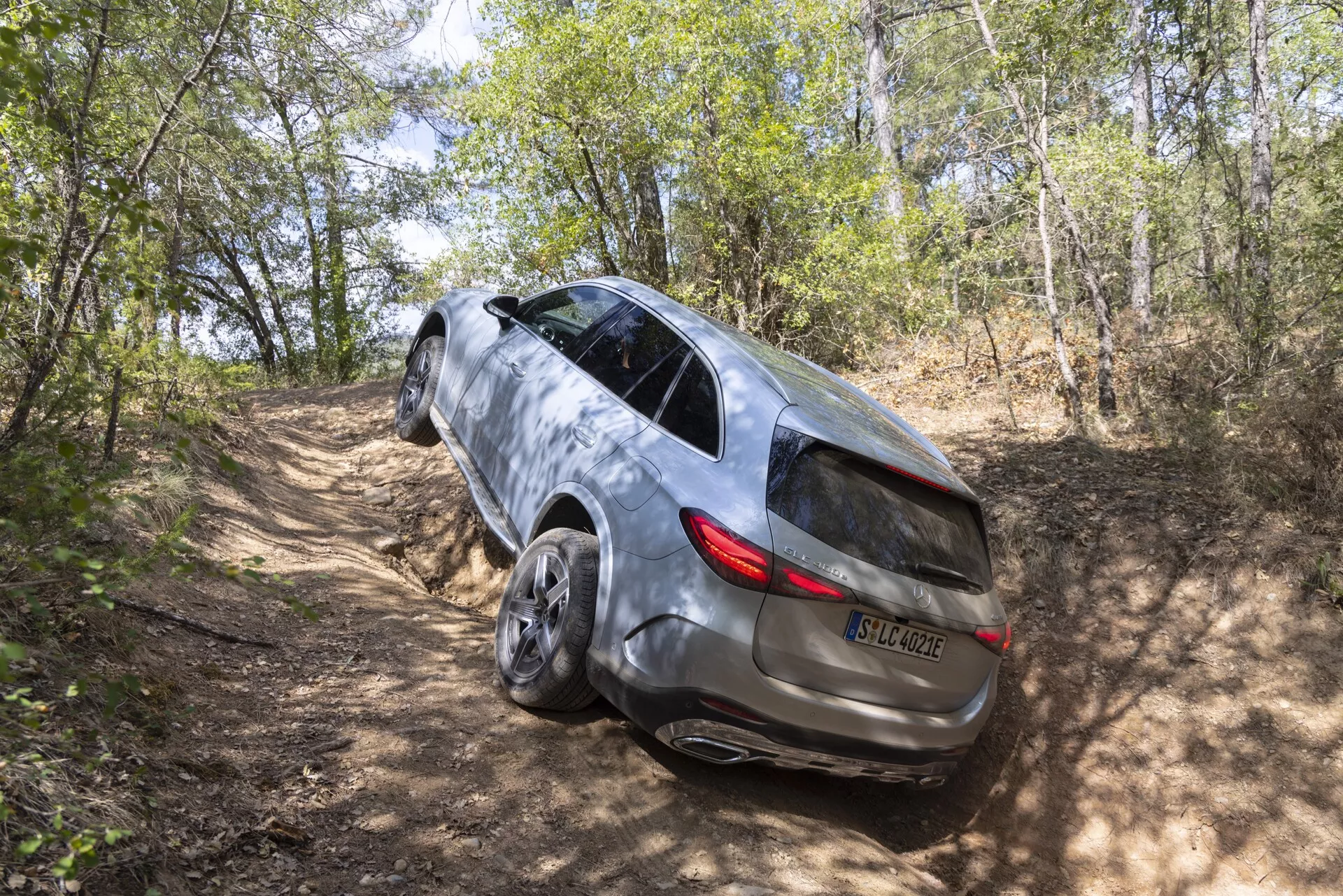 Hög bakre trekvartsvy av en 2022 Mercedes GLC SUV på en ojämn grusväg i ett skogsområde, som visar dess kapacitet för fyrhjulsdrift, elegant design med panoramatak, LED-bakljus, metallic-lack, tonade sidorutor med integrerade blinkers, infällda dörrhandtag och stora lättmetallfälgar.