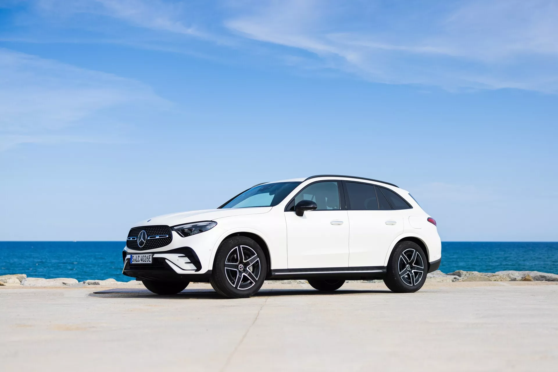 Sidoprofil vy av en modern vit Mercedes GLC 2022 SUV, parkerad mot en naturskön bakgrund med klarblå himmel och lugn horisont vid havet. Fordonet, framträdande centrerat i ramen, visar eleganta, strömlinjeformade kurvor, flerspakiga lättmetallhjul och LED-strålkastare. Den främre grillen och framhjulsdesignen är subtilt framhävda, vilket ger en blandning av lyx och sportighet i en lugn miljö.