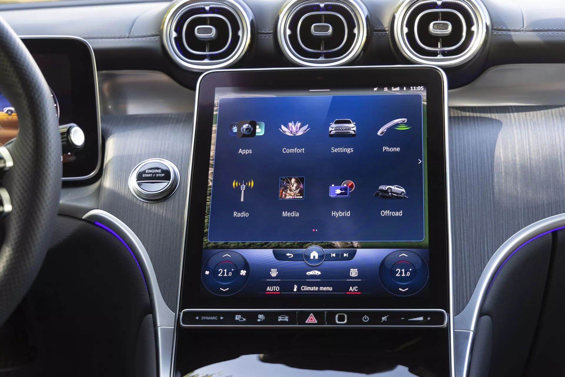 Den centrala konsolen av en Mercedes-Benz GLC 2022, sedd från förarsätet, med fokus på en stor vertikalt placerad pekskärm för infotainmentsystemet som visar alternativ som 'Appar,' 'Komfort,' 'Inställningar,' tillsammans med dubbelzonsklimatanläggning, metalliska luftventiler, start/stopp-knapp och en premiumblandning av borstad metall och högglansigt svart finish.